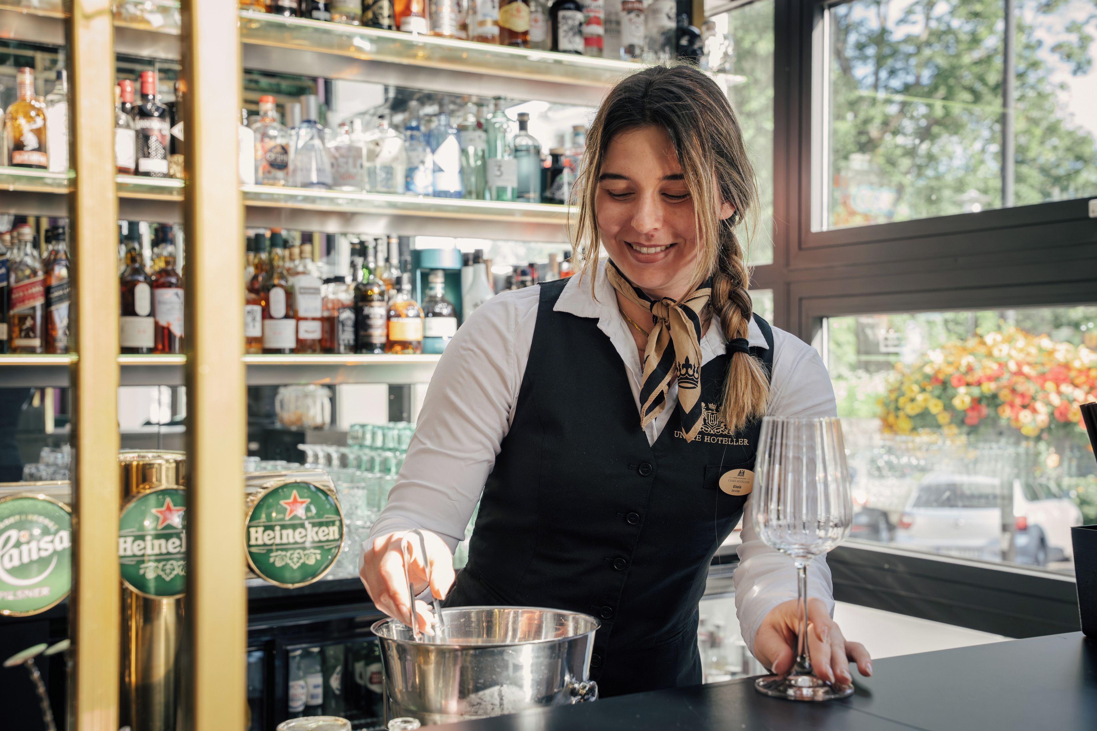 A bartender preparing a drink behind a bar with various bottles and a window view.