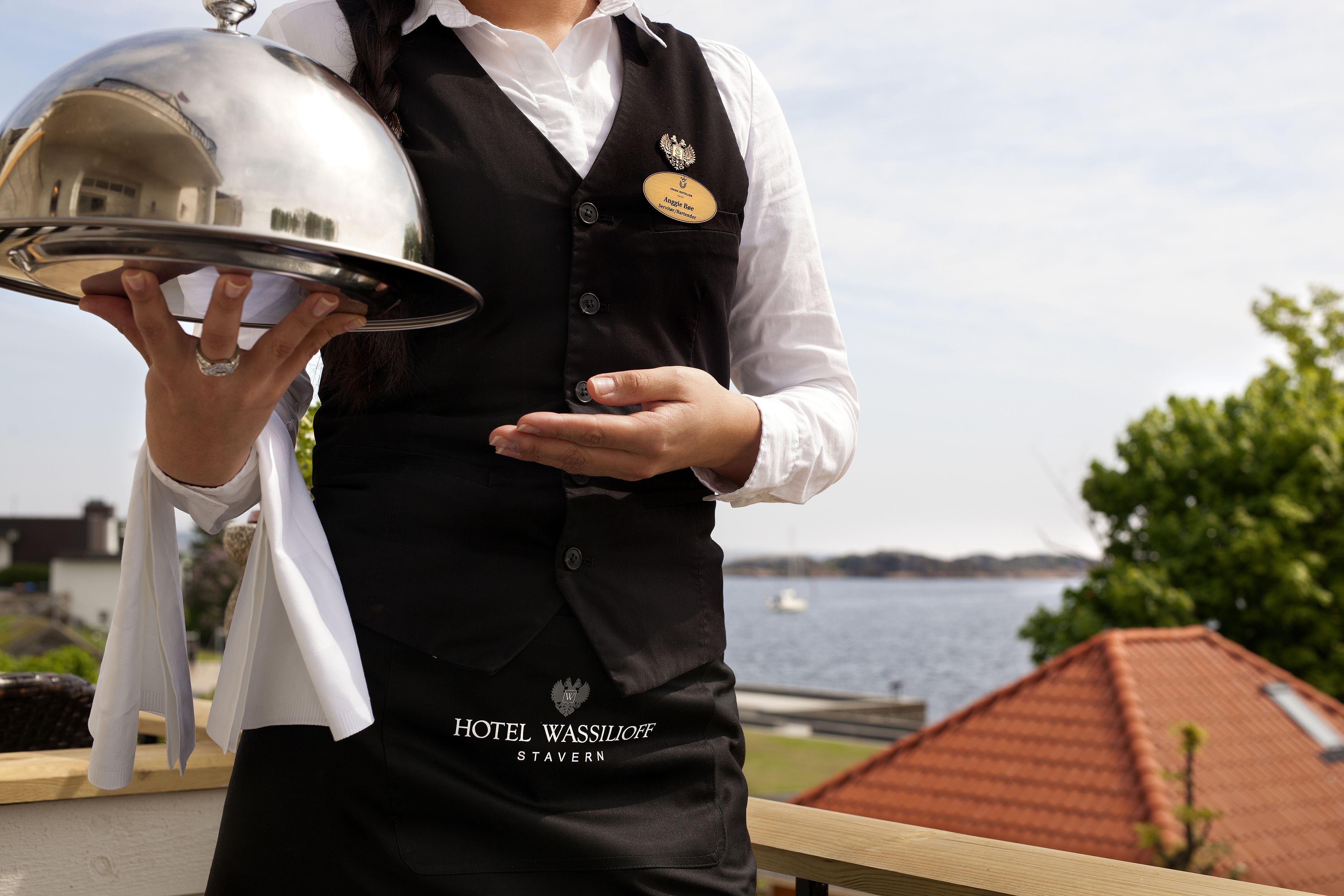 Una persona con chaleco negro, placa de identificación y logotipo de hotel, sosteniendo una bandeja plateada.