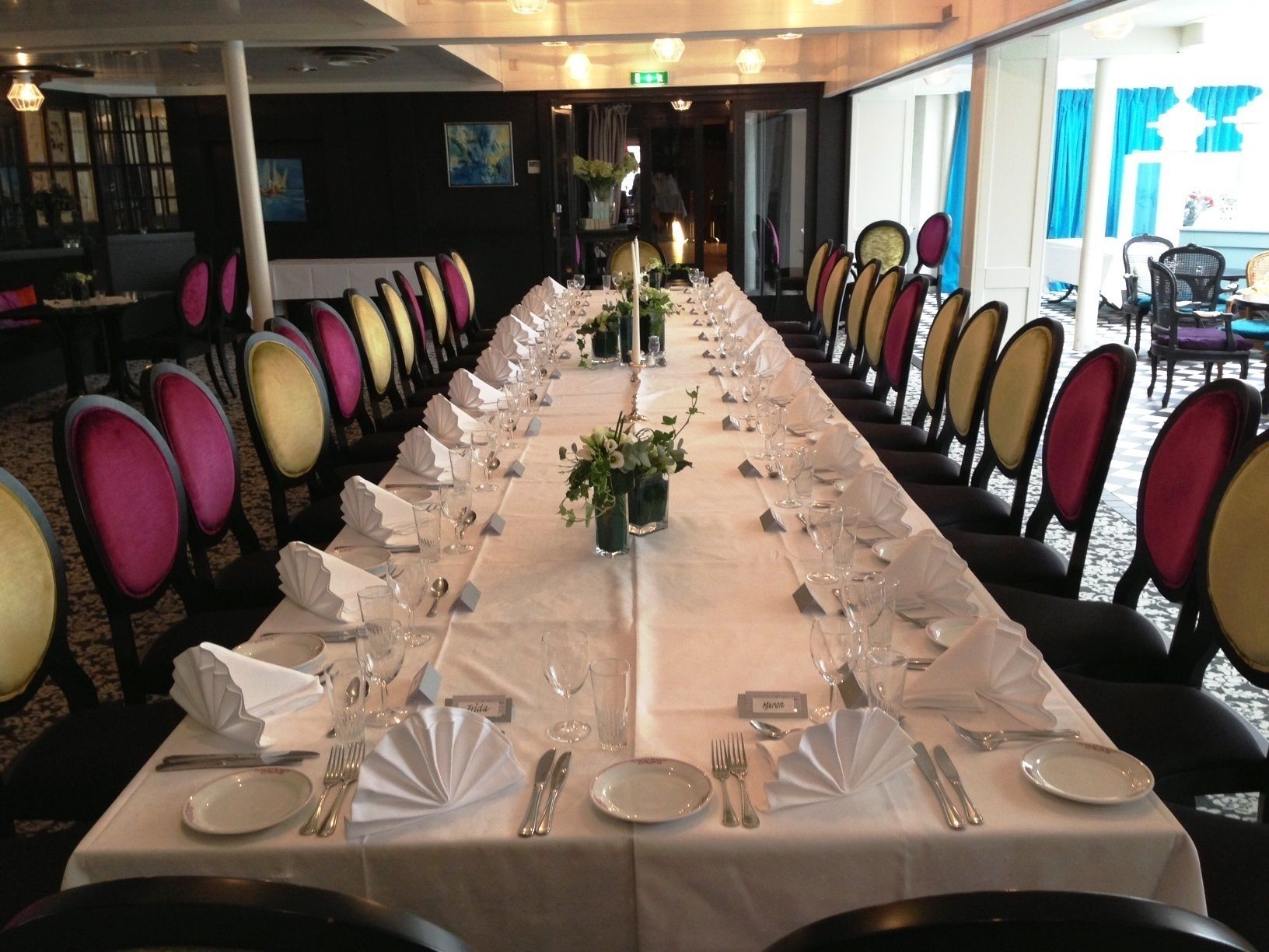 Una larga mesa de banquete dispuesta con manteles blancos, sillas con cojines de colores y un jarrón con flores.