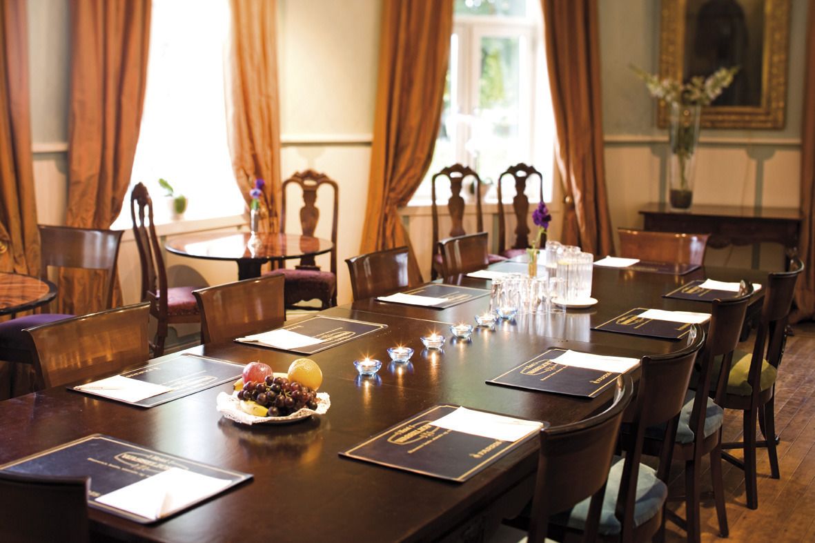 Une salle à manger élégante, avec une longue table dressée pour une réunion, des chaises en bois et un vase rempli de fleurs placé sur une table d’appoint.