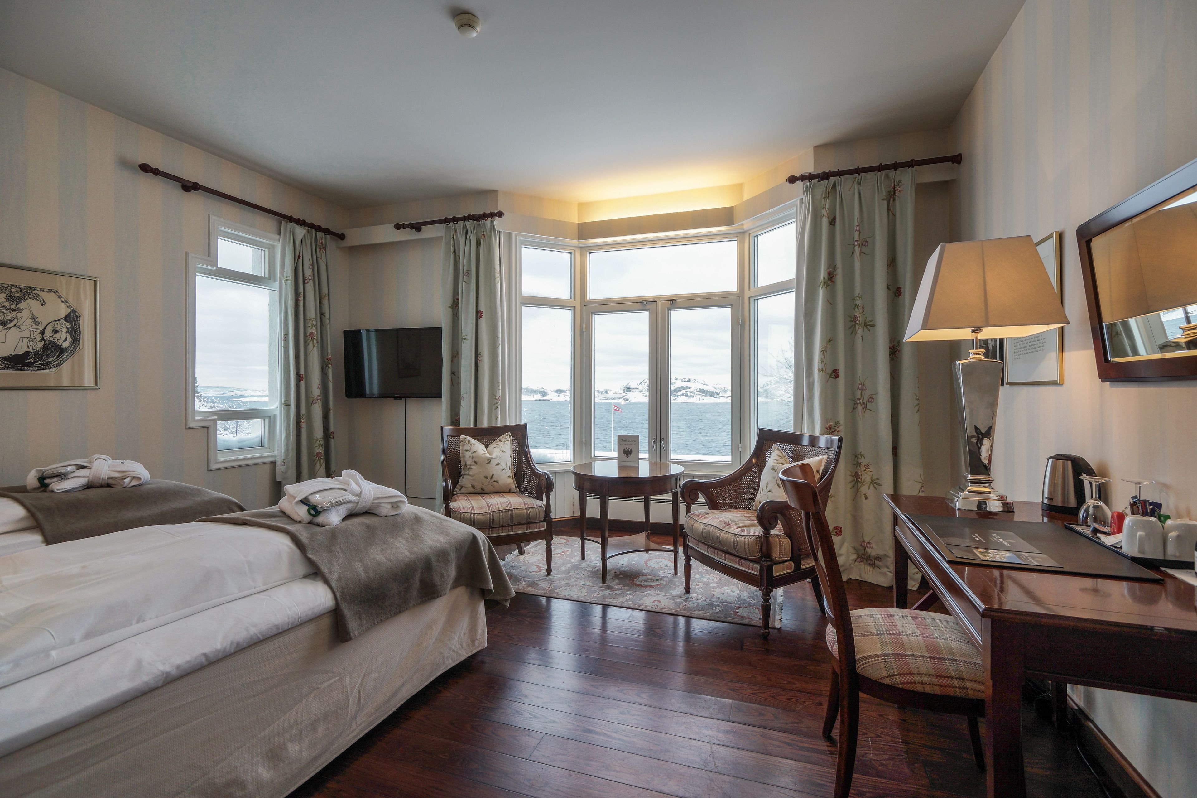 A hotel room with a bed, two chairs, a table, a desk, a lamp, a television, and a large window with curtains.