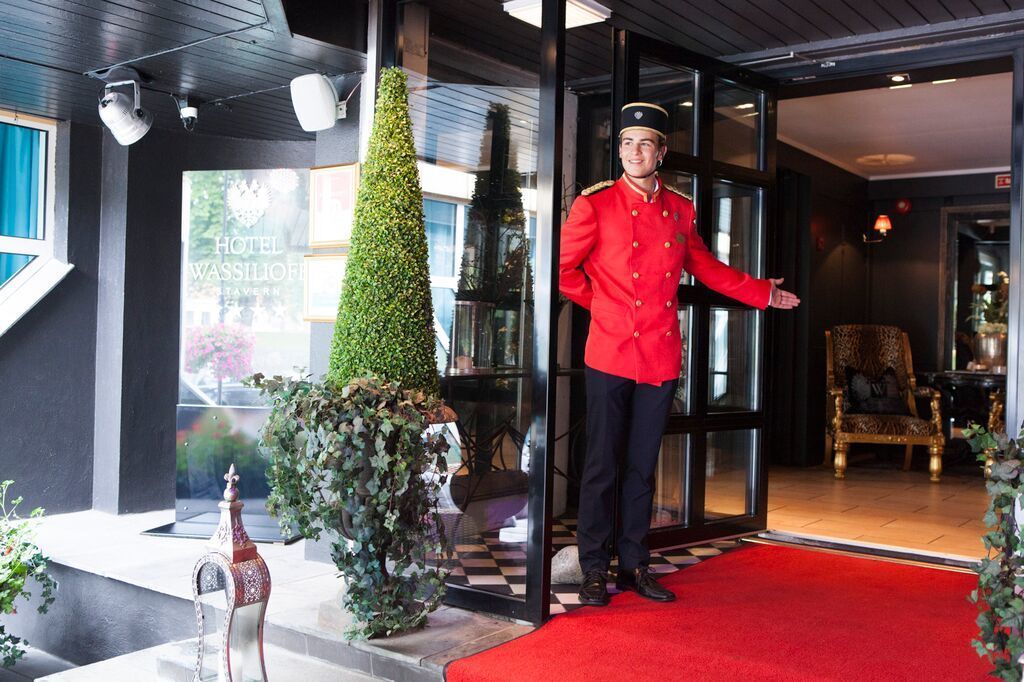 A hotel entrance with a red carpet, a uniformed staff member, and a large potted tree.