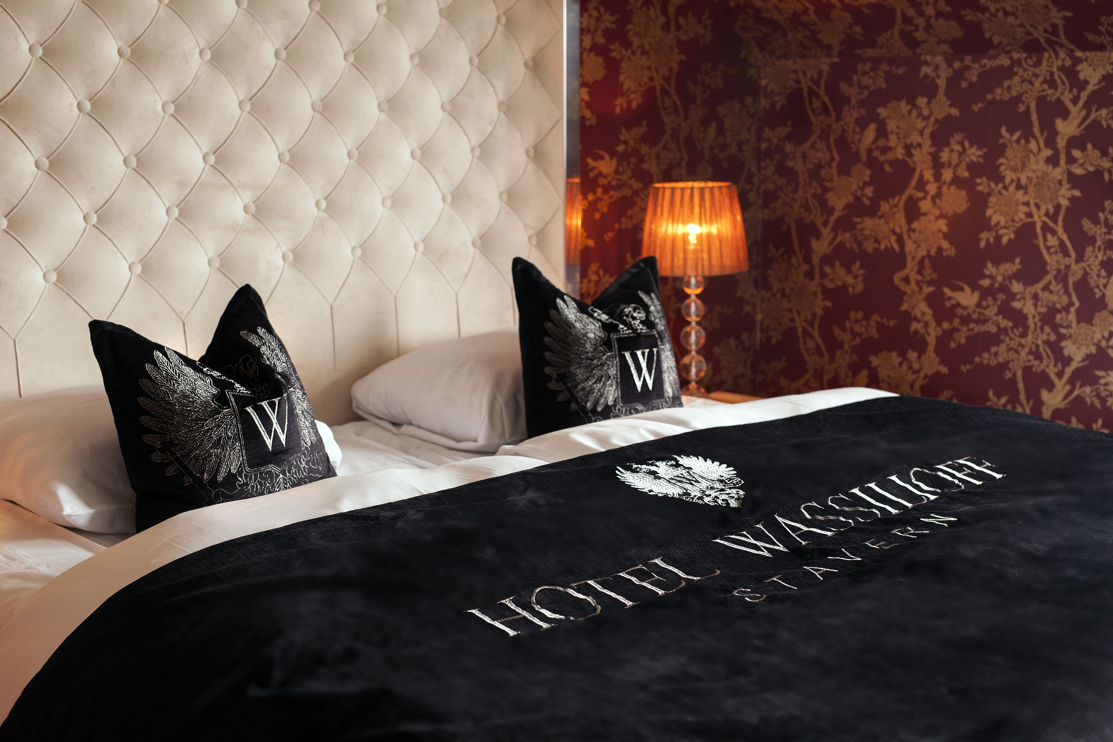 Una habitación de hotel con cabecera acolchada, colcha negra con texto bordado y una lámpara decorativa.