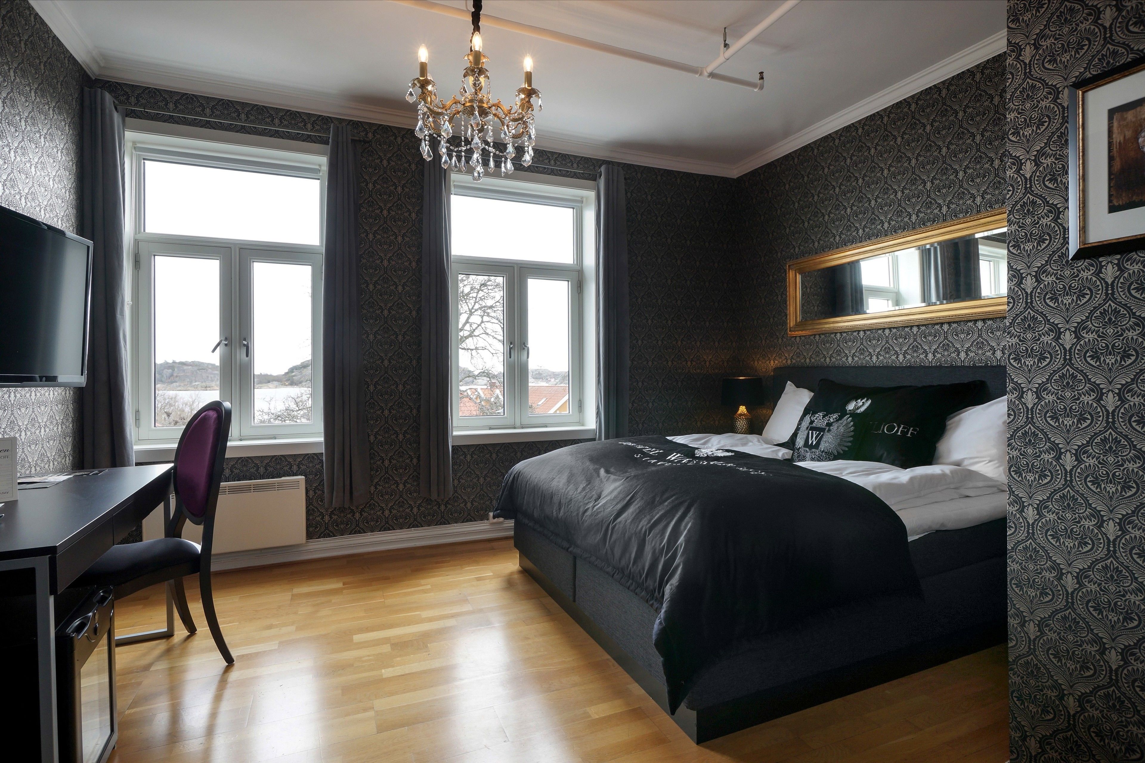 A bedroom with a large bed, a desk, and a chair. There is a chandelier, a window, and a mirror on the wall.