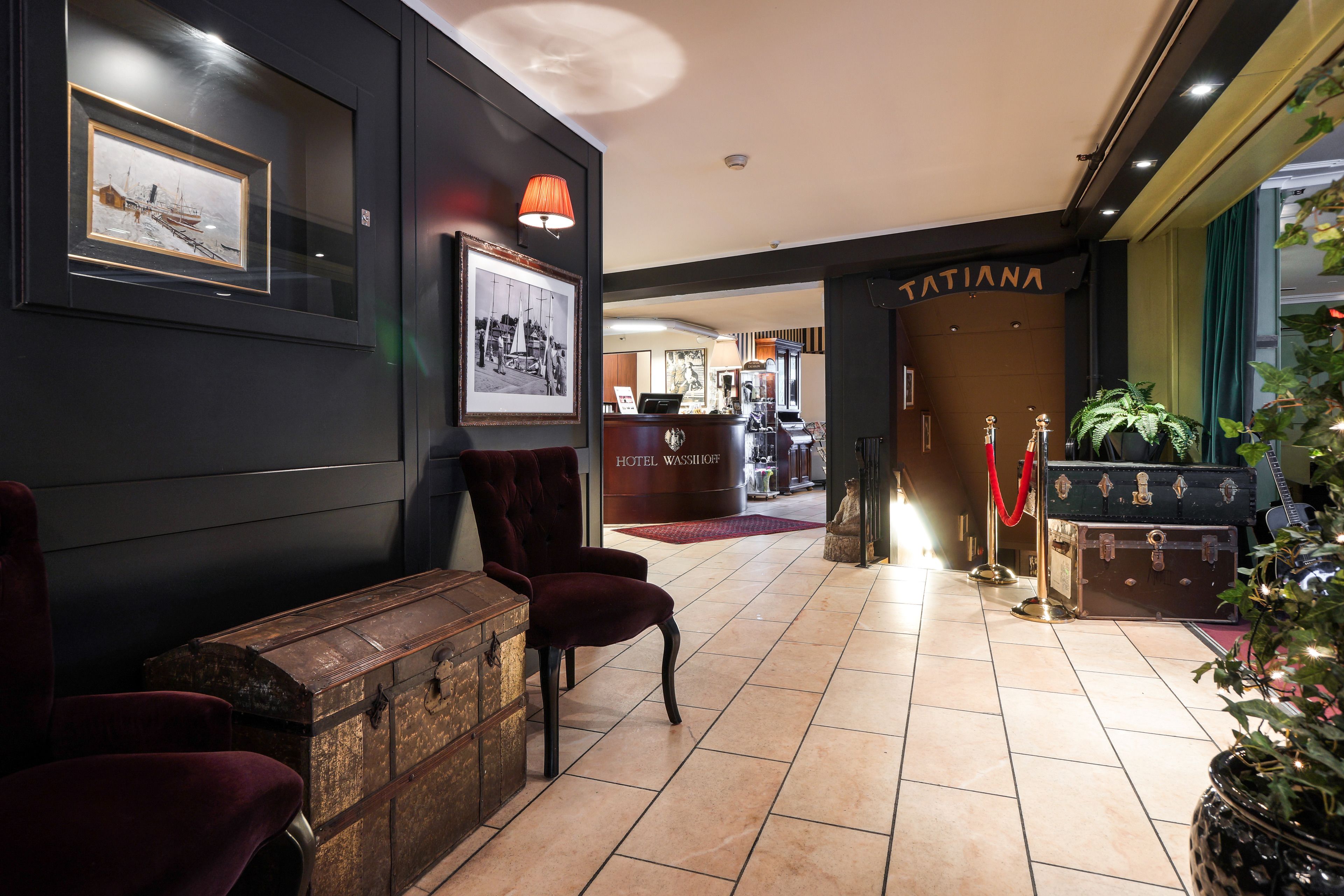 A lobby with a reception desk, framed artwork on the wall, a red velvet chair, a vintage trunk, and a potted plant.