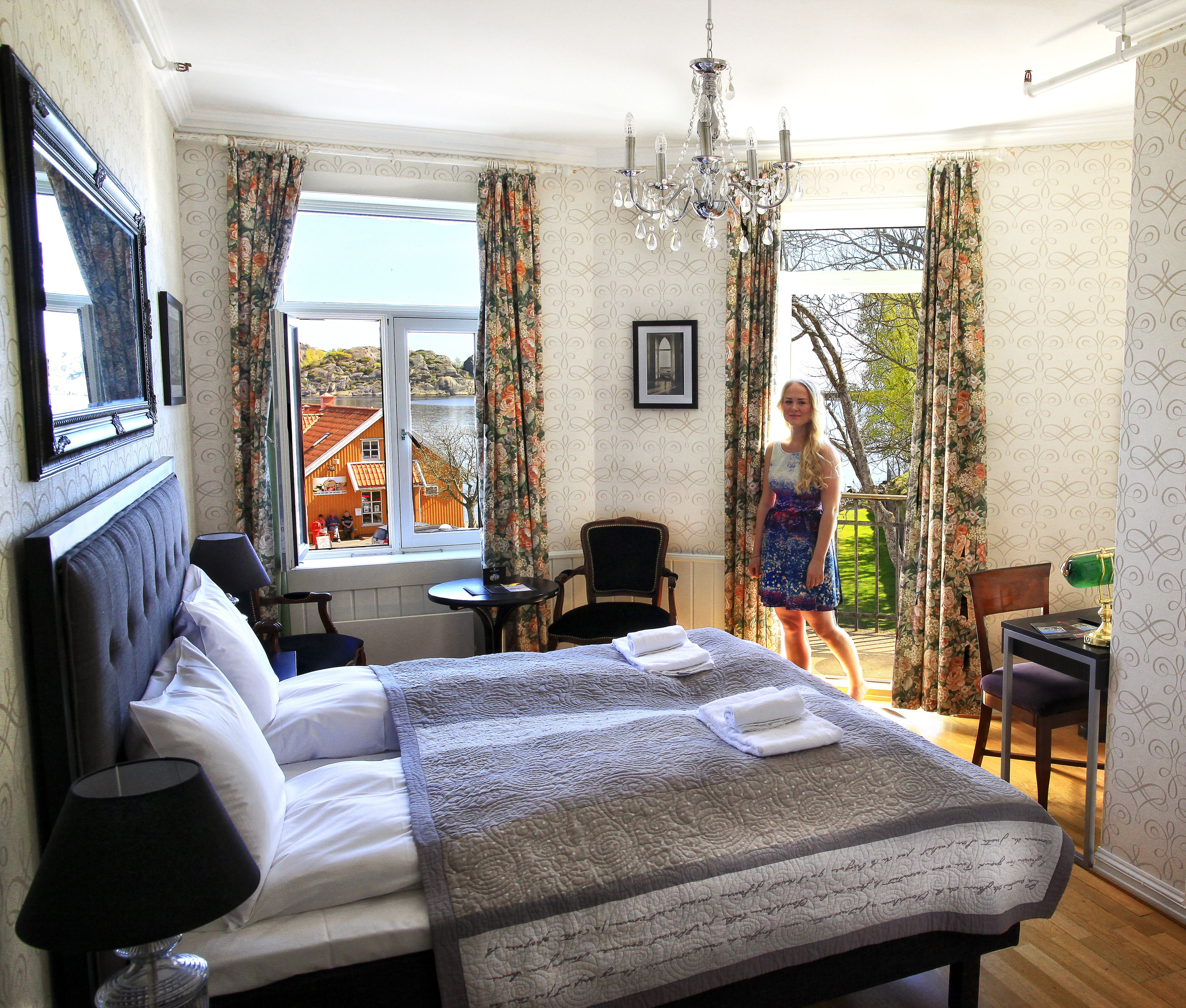 A bedroom with a large bed, a desk, and a chair. There is a window with curtains, a chandelier, and a view of buildings outside.