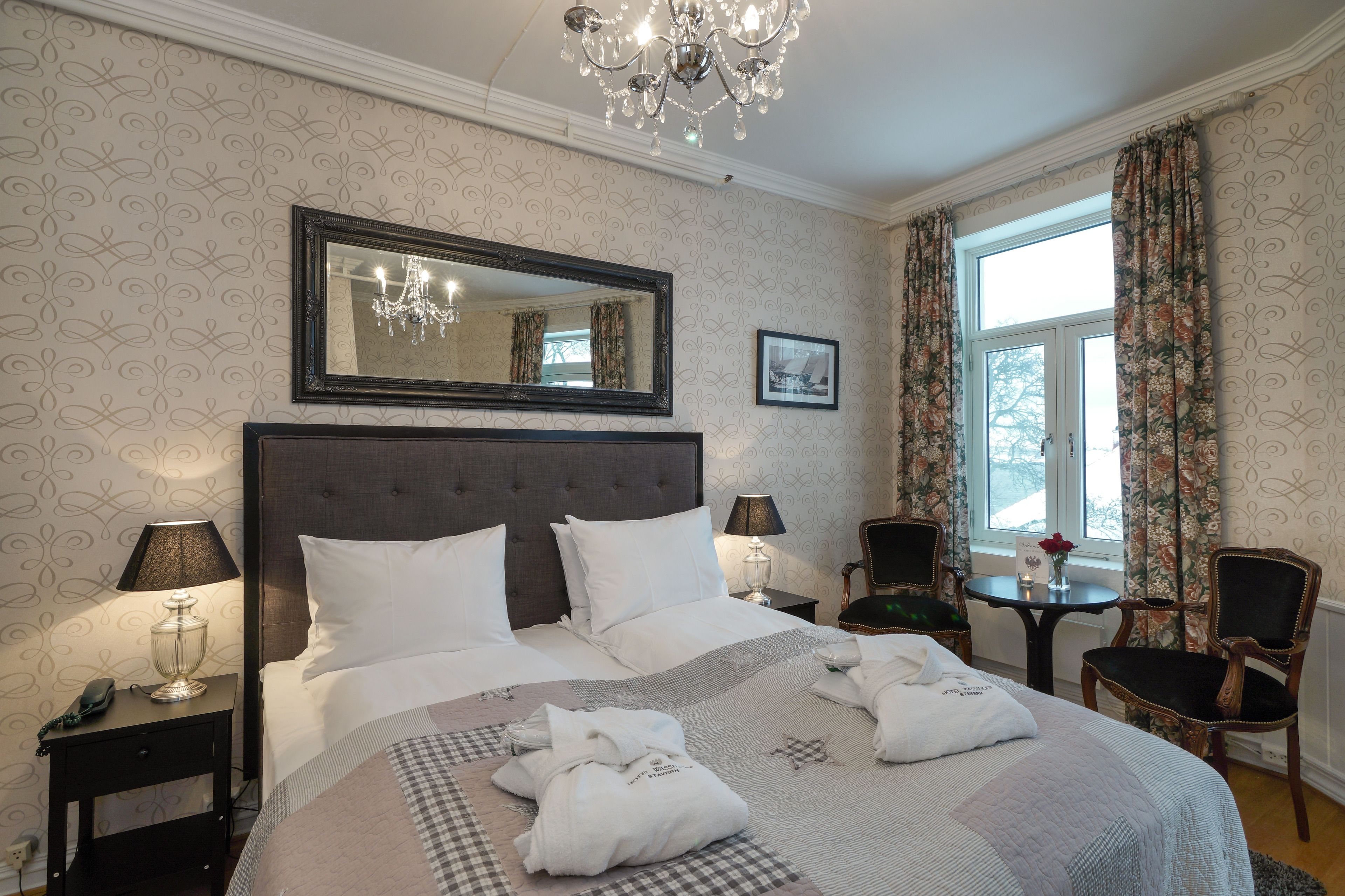 Habitación de hotel con una cama grande, dos mesitas de noche con lámparas, un candelabro, una ventana con cortinas y una silla.