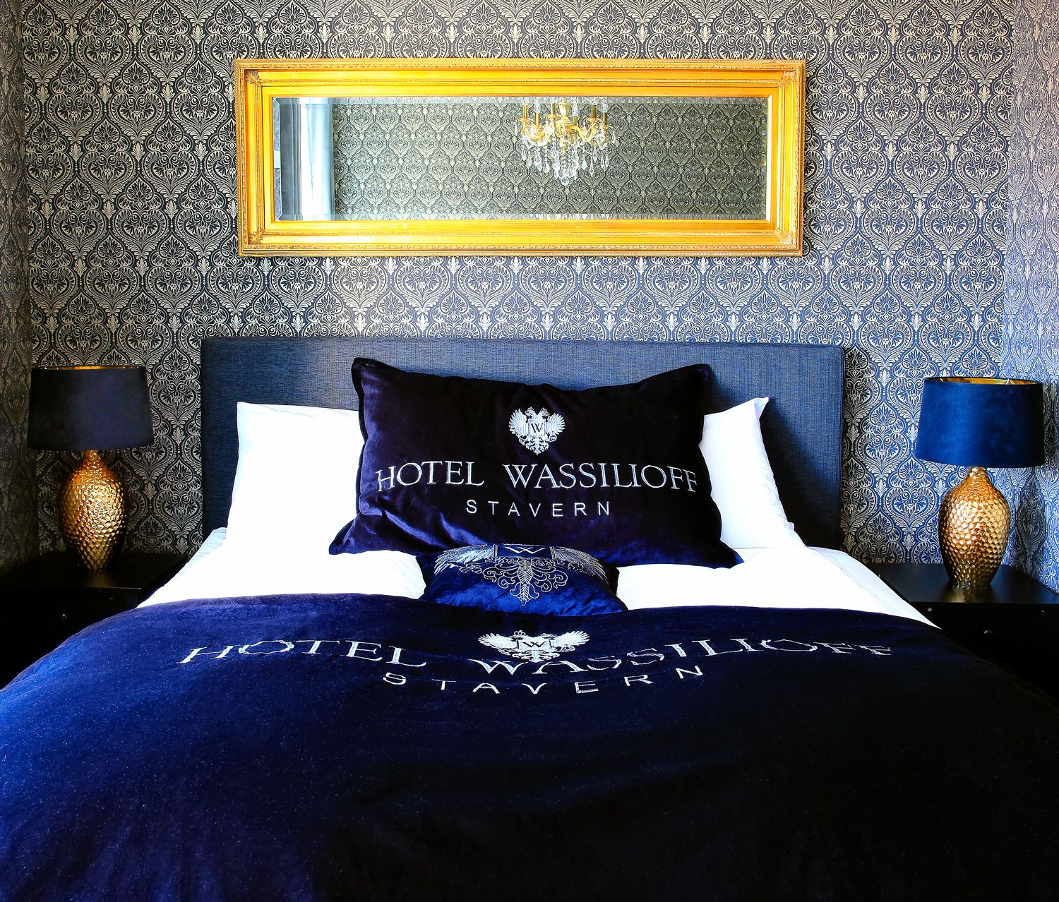 A hotel room with a patterned wallpaper, a bed with a dark blue bedspread and pillows, a gold-framed mirror above the bed, and two lamps on either side of the bed.