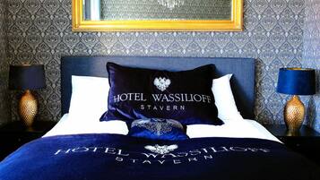 A hotel room with a patterned wallpaper, a bed with a dark blue bedspread and pillows, a gold-framed mirror above the bed, and two lamps on either side of the bed.