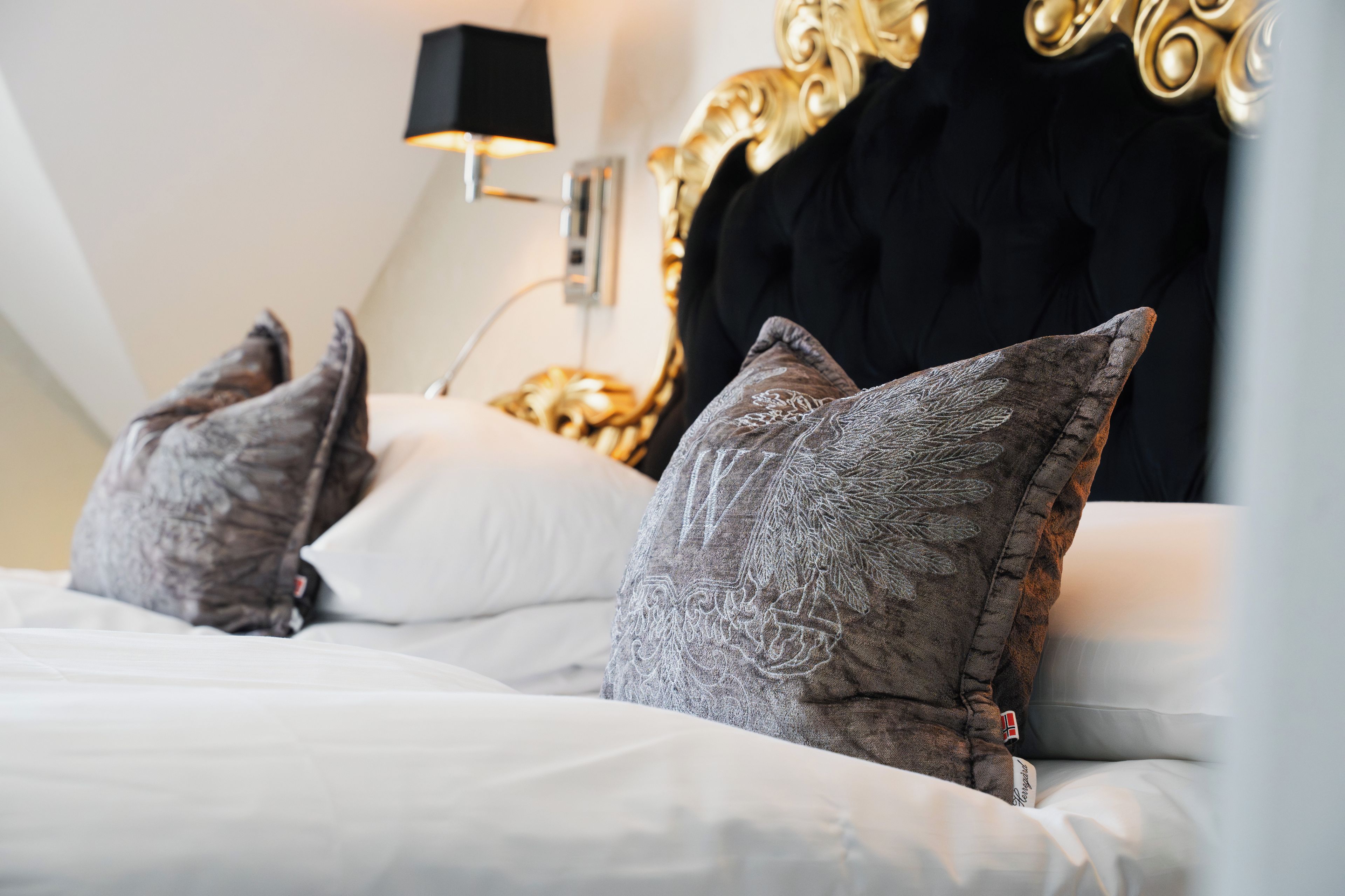 A bed with white bedding and grey pillows, a black lamp on a bedside table, and a gold-framed headboard.