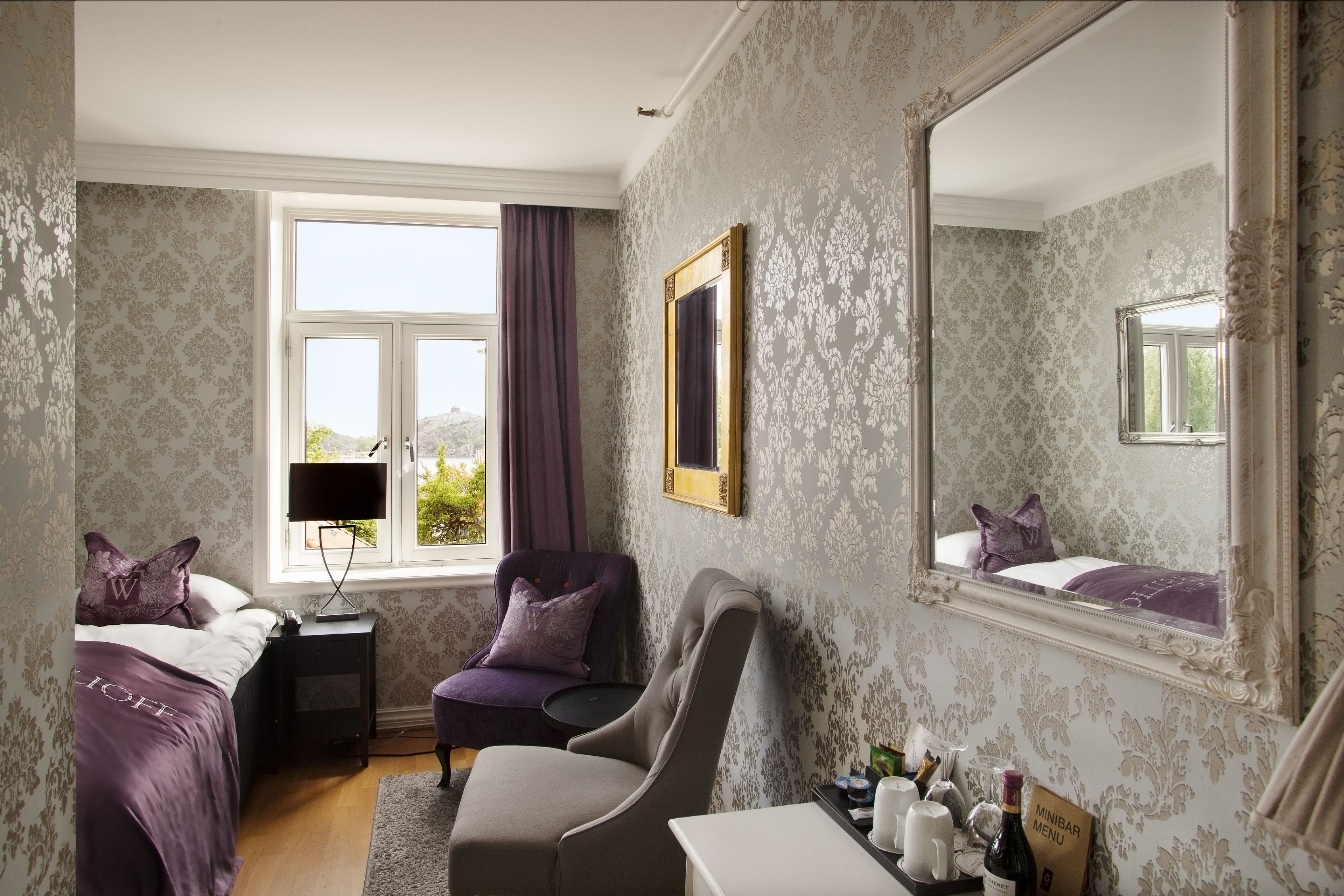 A hotel room with a large mirror, a bed with purple bedding, a chair, a small table with a minibar, and a window with curtains.