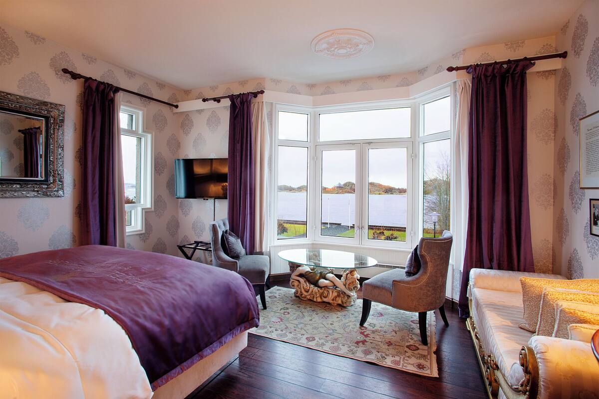 A hotel room with a large bed, a sofa, a chair, a small table, and a bay window with curtains.