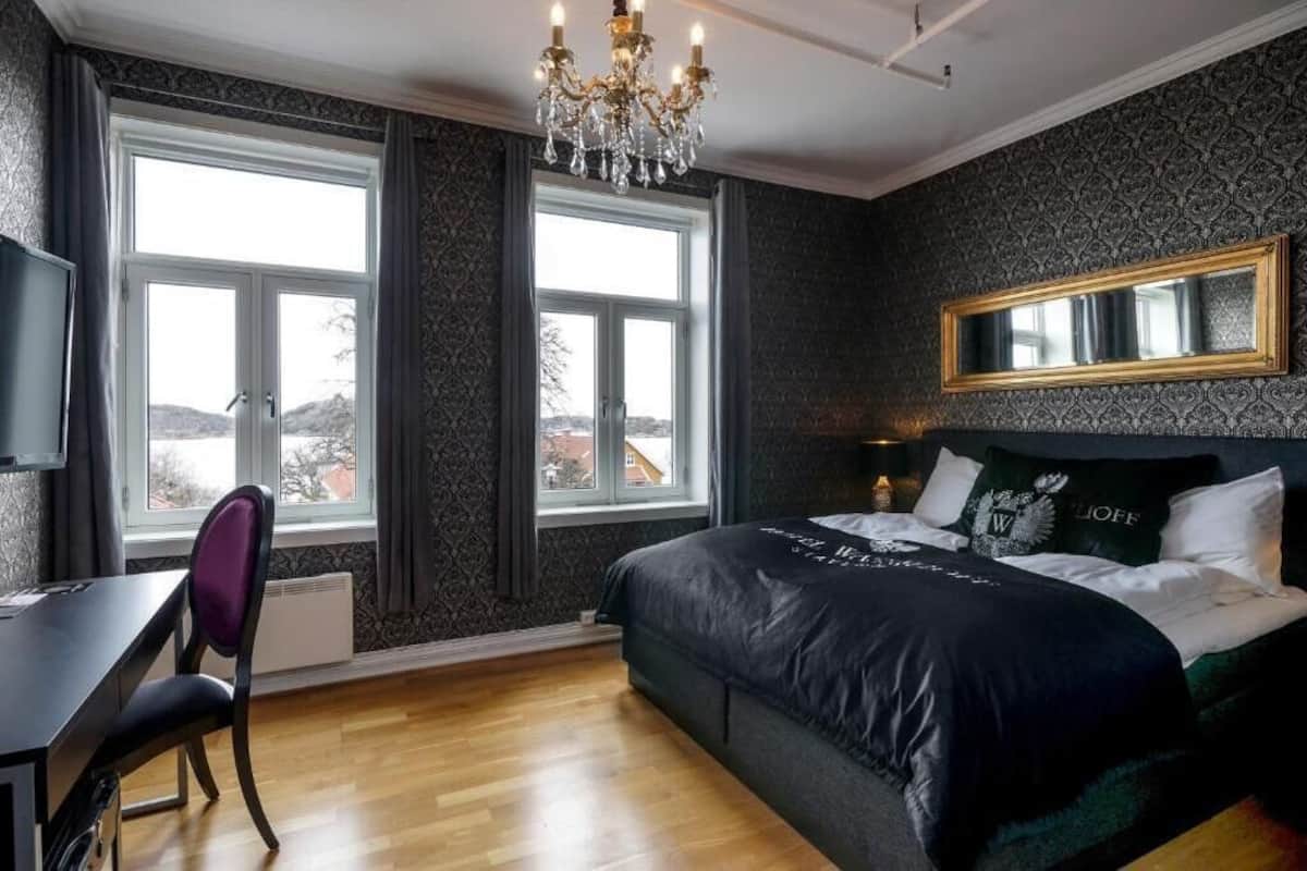 A bedroom with a large bed, a desk, and a chair. There is a window with a view of the outside.