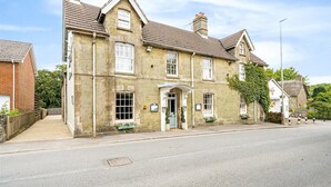 A two-story stone building with a traditional design, featuring a central entrance with a small porch, multiple windows, and a brick wall adjacent to a paved street.
