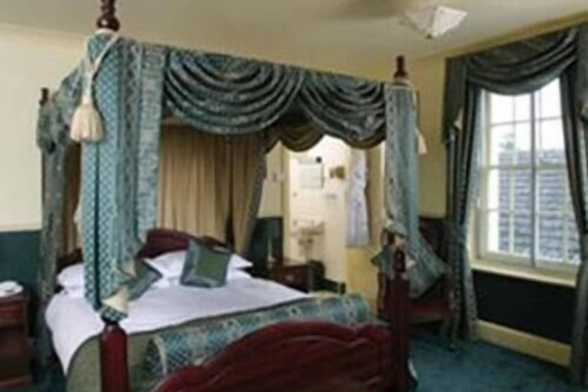 Una cama con dosel sobre cuatro columnas, con cabecera tapizada de rojo, una mesita de noche de madera y una ventana con cortinas.