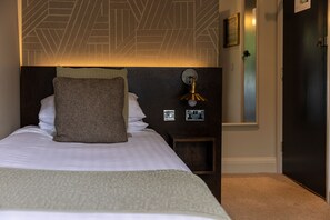 A neatly made bed with pillows and a blanket, a wall-mounted light fixture, and a door. - The Royal Hotel (Carnforth)