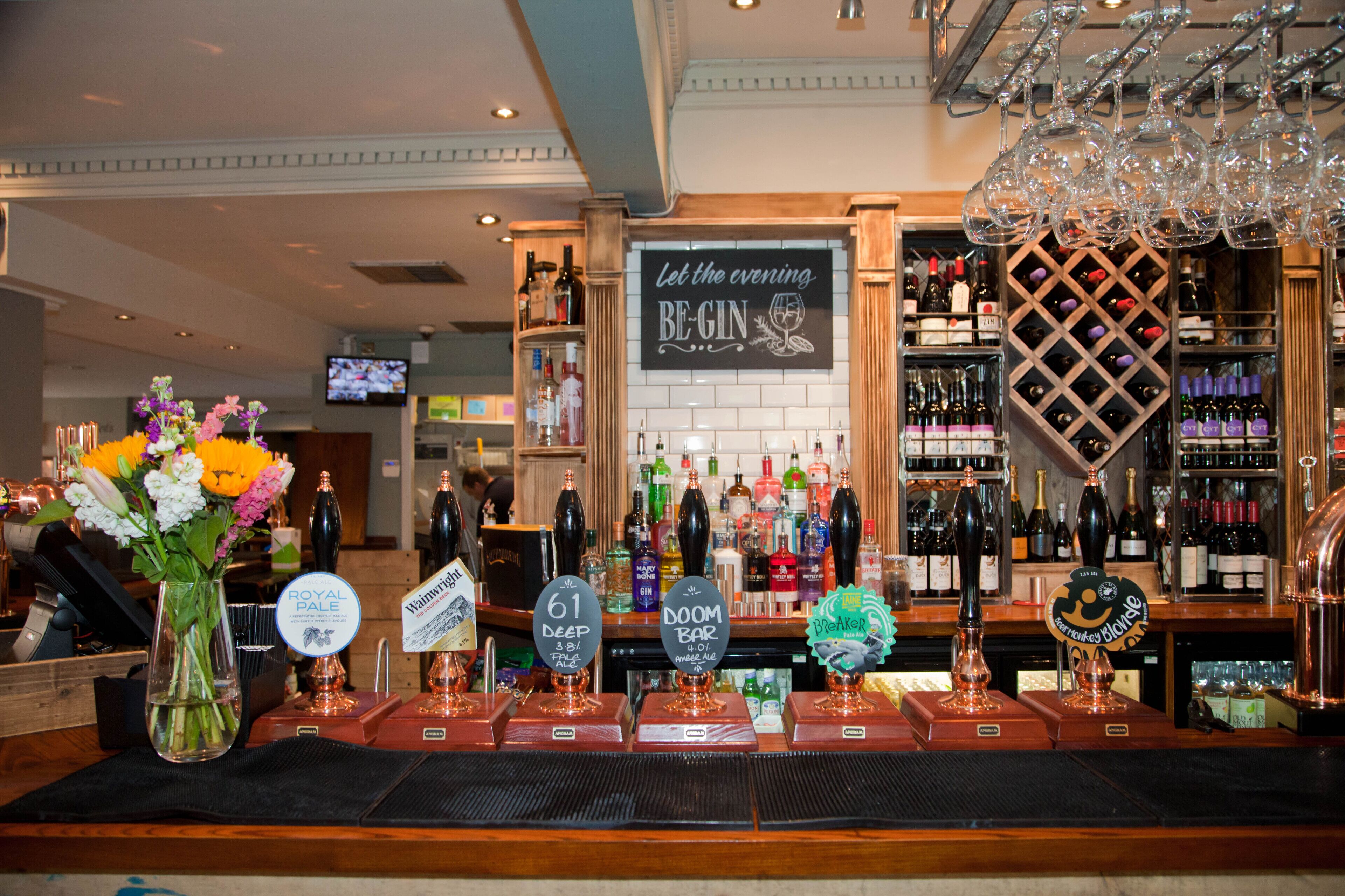 A bar area with a variety of bottles and glasses, a sign that reads "Let the evening BE GIN," and a television in the background.