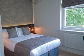A neatly made bed with a headboard, a bedside table with a lamp, and a window with a view of trees. - The Royal Hotel (Carnforth)