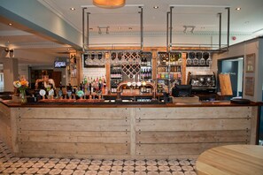 Uma área de bar com balcões de madeira, diversos tipos de garrafas, copos e uma tela de televisão.