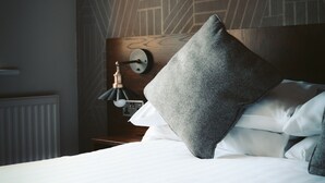 A neatly made bed with white linens and a grey pillow, a wall clock, and a modern light fixture.