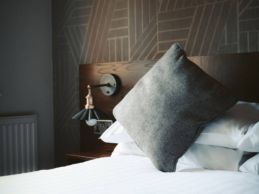 A neatly made bed with white linens and a grey pillow, a wall clock, and a modern light fixture.