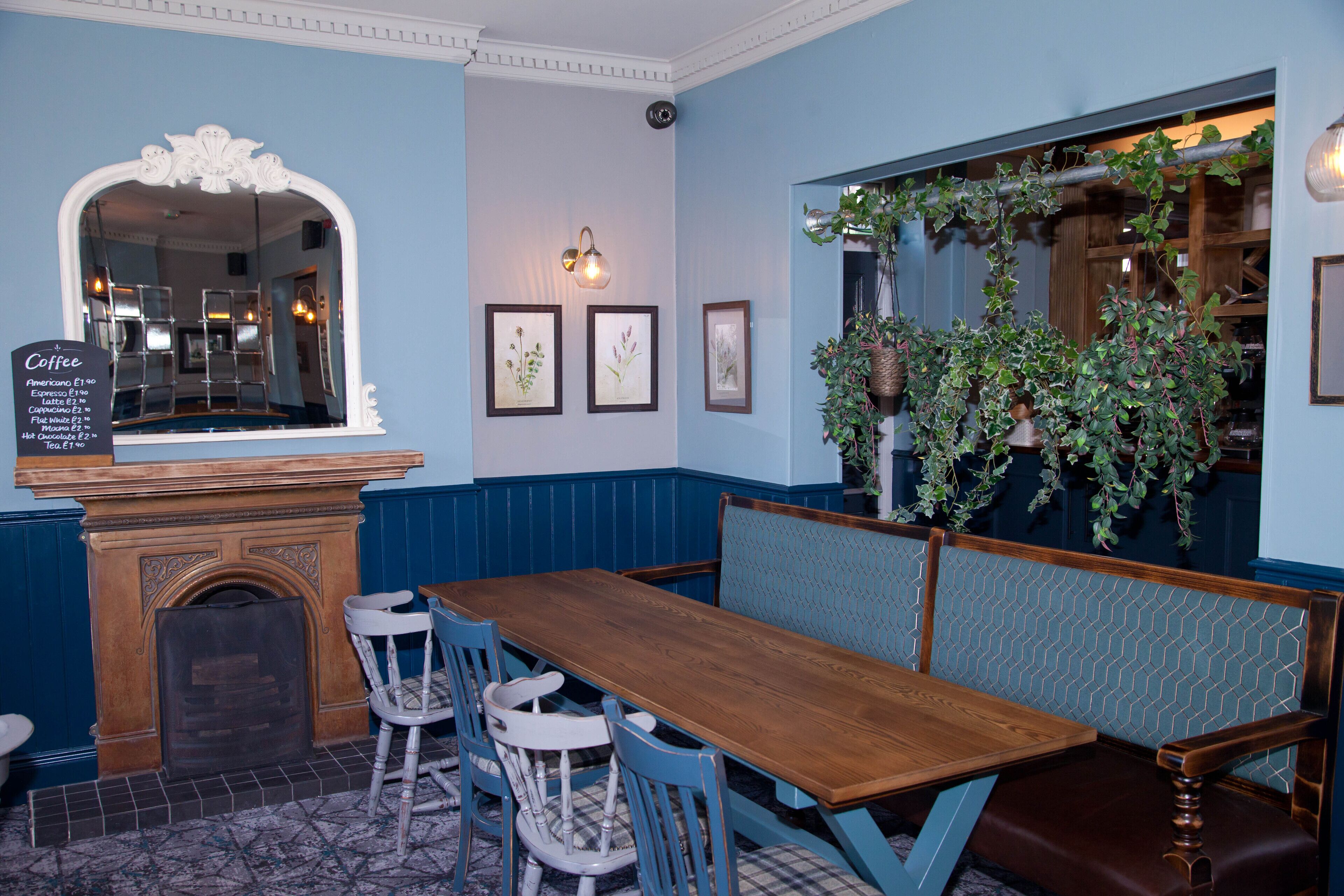 Un restaurant avec une table en bois, des chaises bleues, un banc, une cheminée, des tableaux encadrés et une plante suspendue.