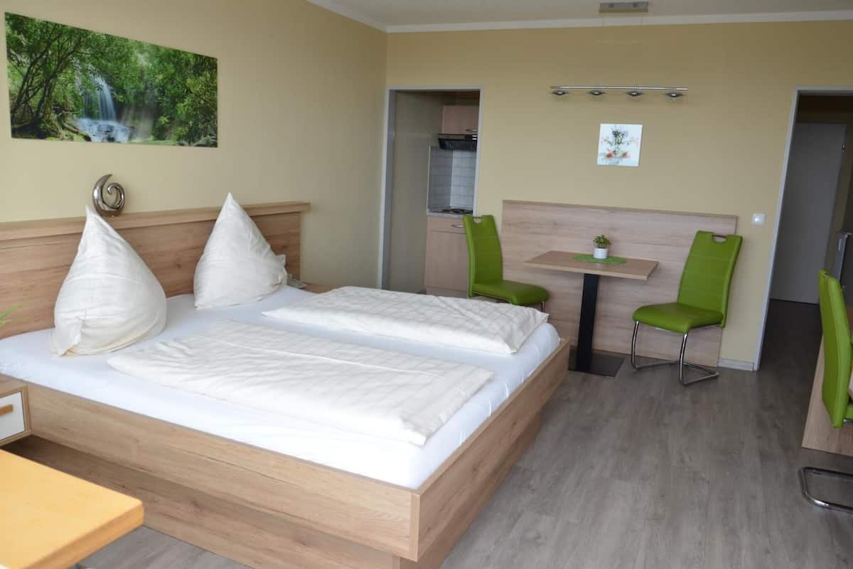 A double bed with white bedding, a wooden headboard, a green chair, a small table with a plant, and a painting of a waterfall on the wall.