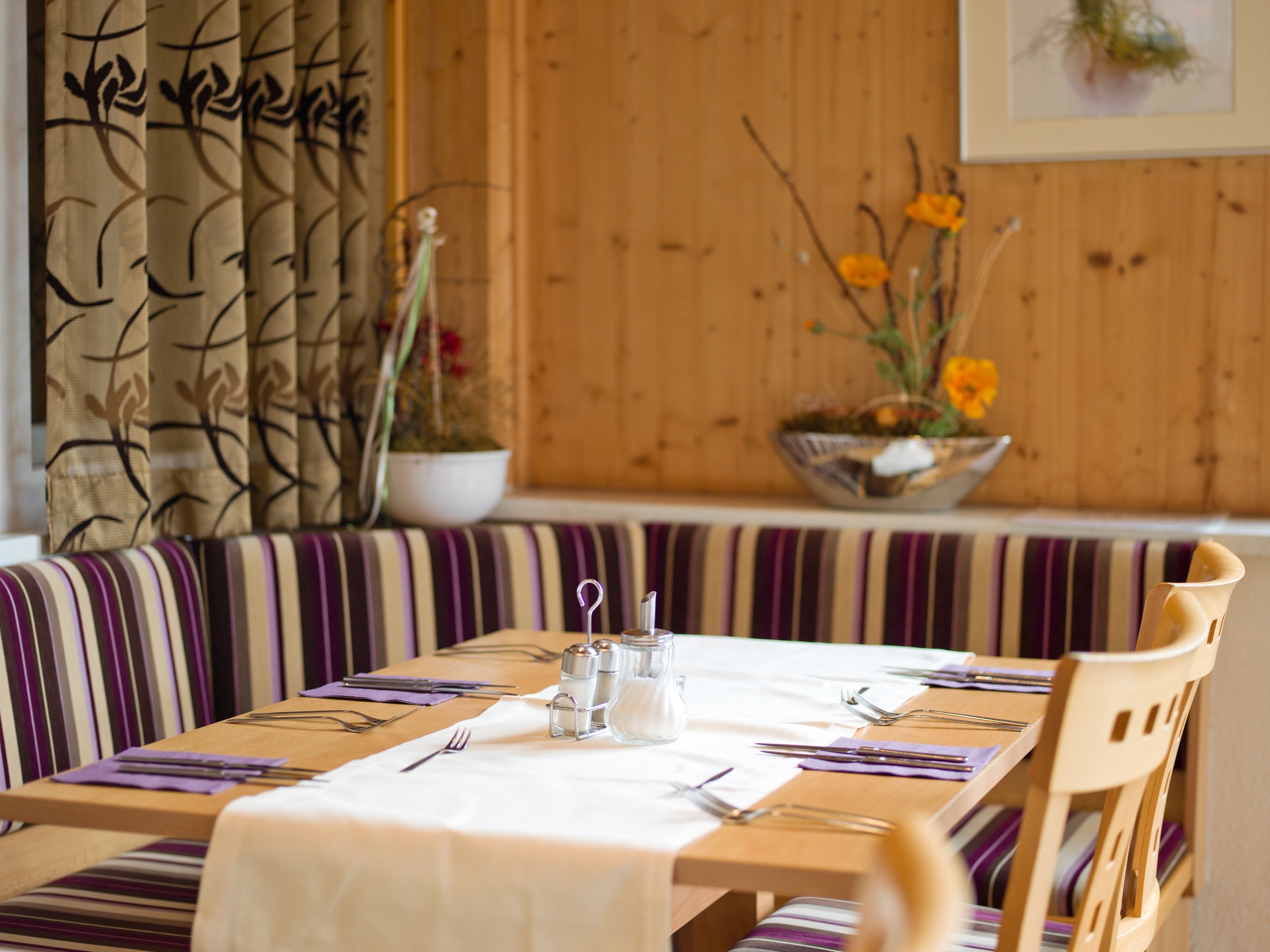 A dining area with a wooden table set for four, striped cushions, and a potted plant with yellow flowers.