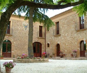 A stone building with multiple arches and balconies, surrounded by potted plants and flowers. - Villa Trigona (Piazza Armerina)