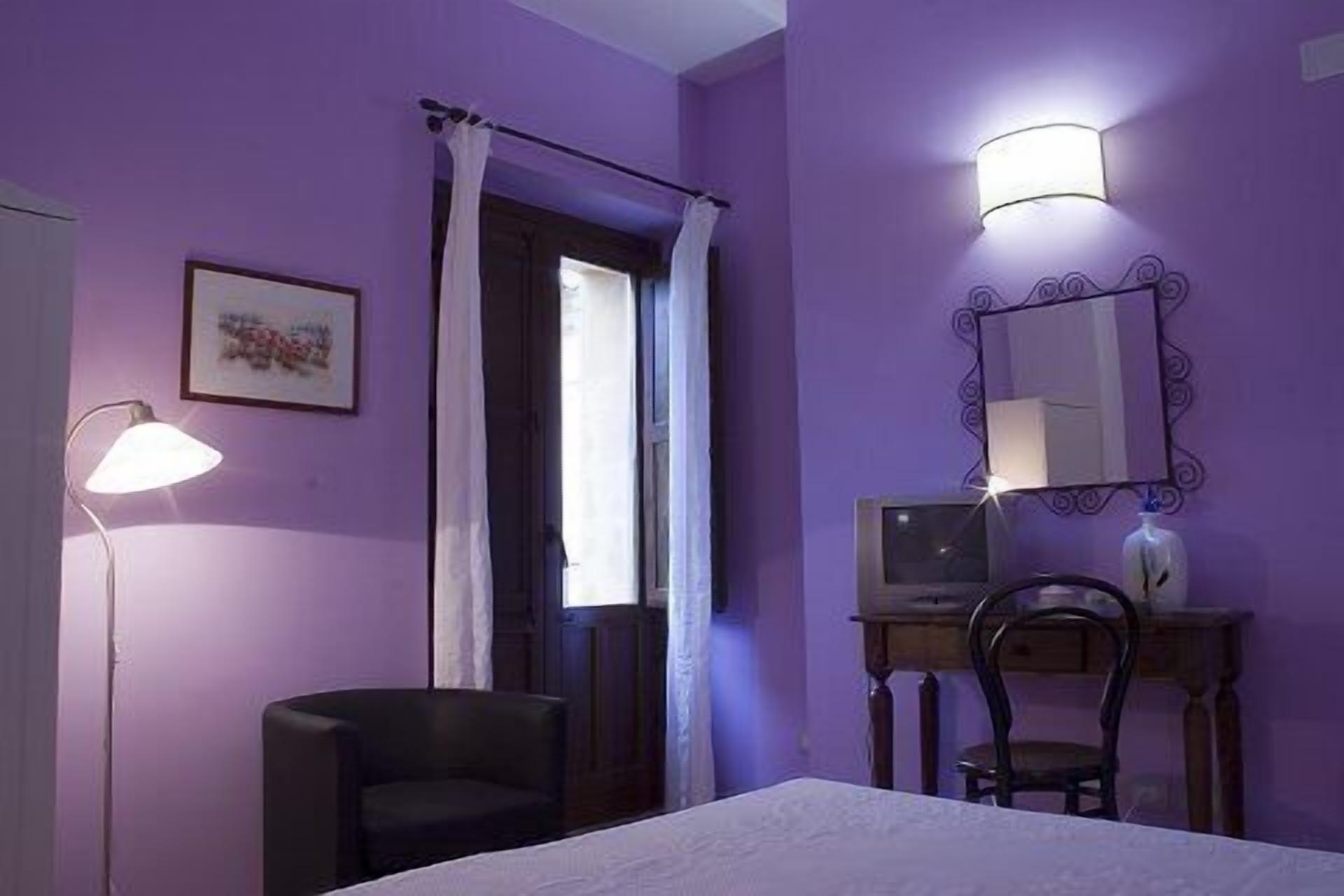 A room with purple walls, a bed, a desk with a chair, a lamp, and a mirror.