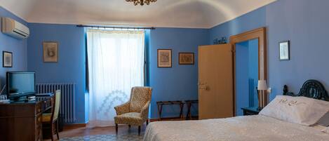 Un dormitorio con cama, una silla, un escritorio con computadora, una ventana con cortinas y un candelabro.