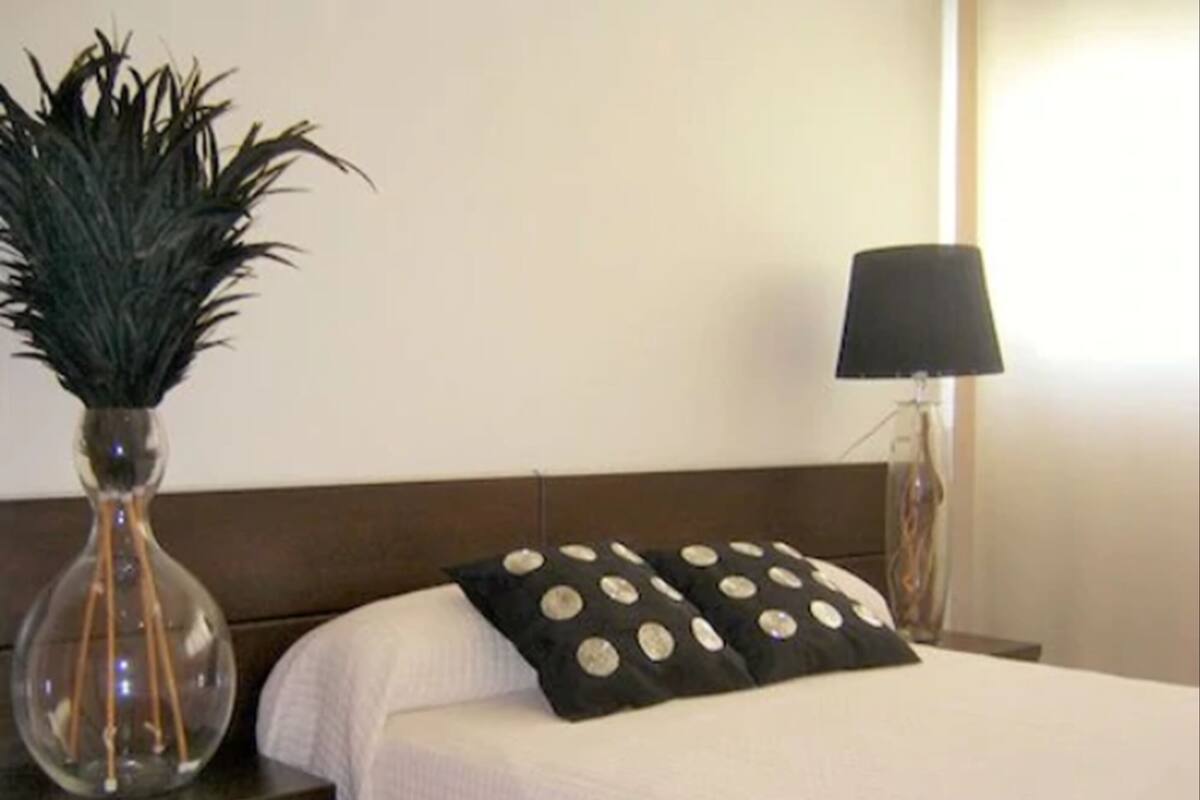 Una cama bien hecha con una colcha blanca y un almohadón negro con diseños circulares. Hay una jarra transparente con una planta sobre una mesita de noche. Una lámpara está en la mesita junto a la cama.
