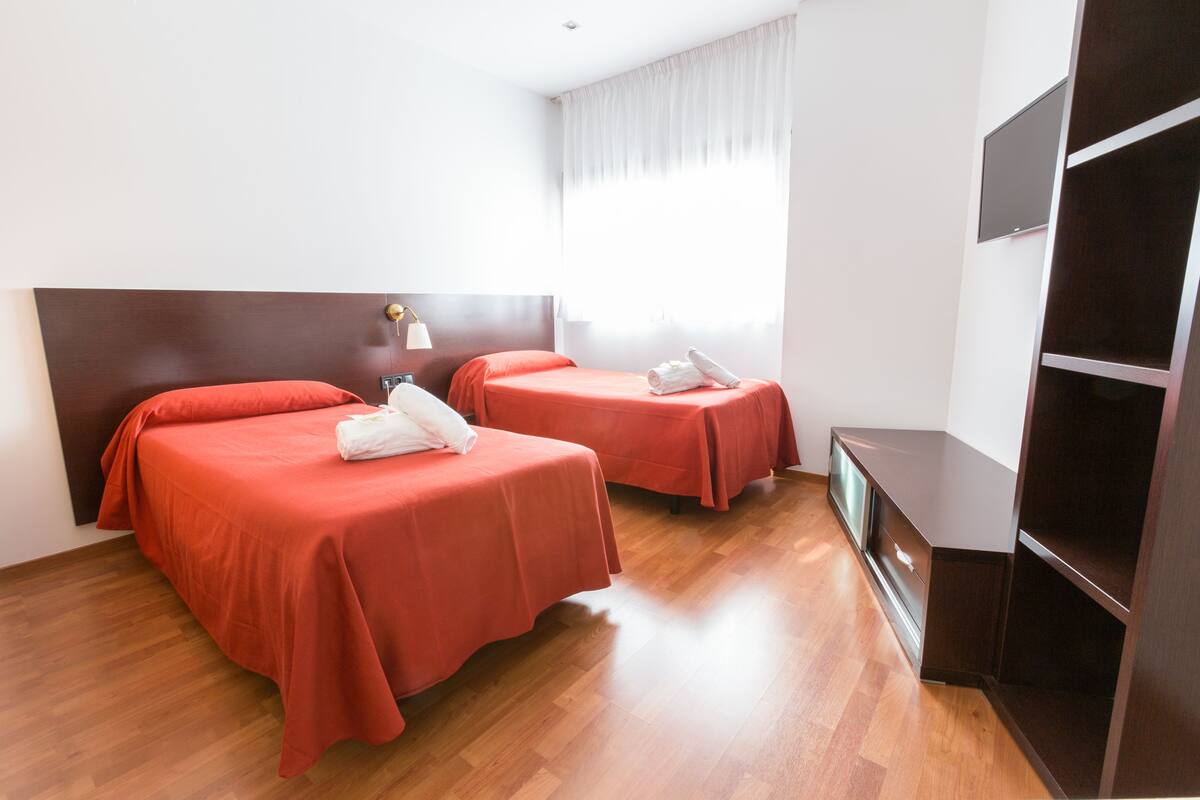 Habitación de hotel con dos camas, piso de madera, televisión y cortina blanca.