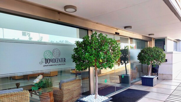 Un bâtiment moderne doté d’une façade en verre, d’un espace extérieur aménagé avec des meubles en osier et de plantes en pot.