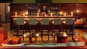 A bar area with a bartender, wooden bar counter, bar stools, and a seating area with a sofa and armchairs. - Penmaenuchaf Hotel (Dolgellau)