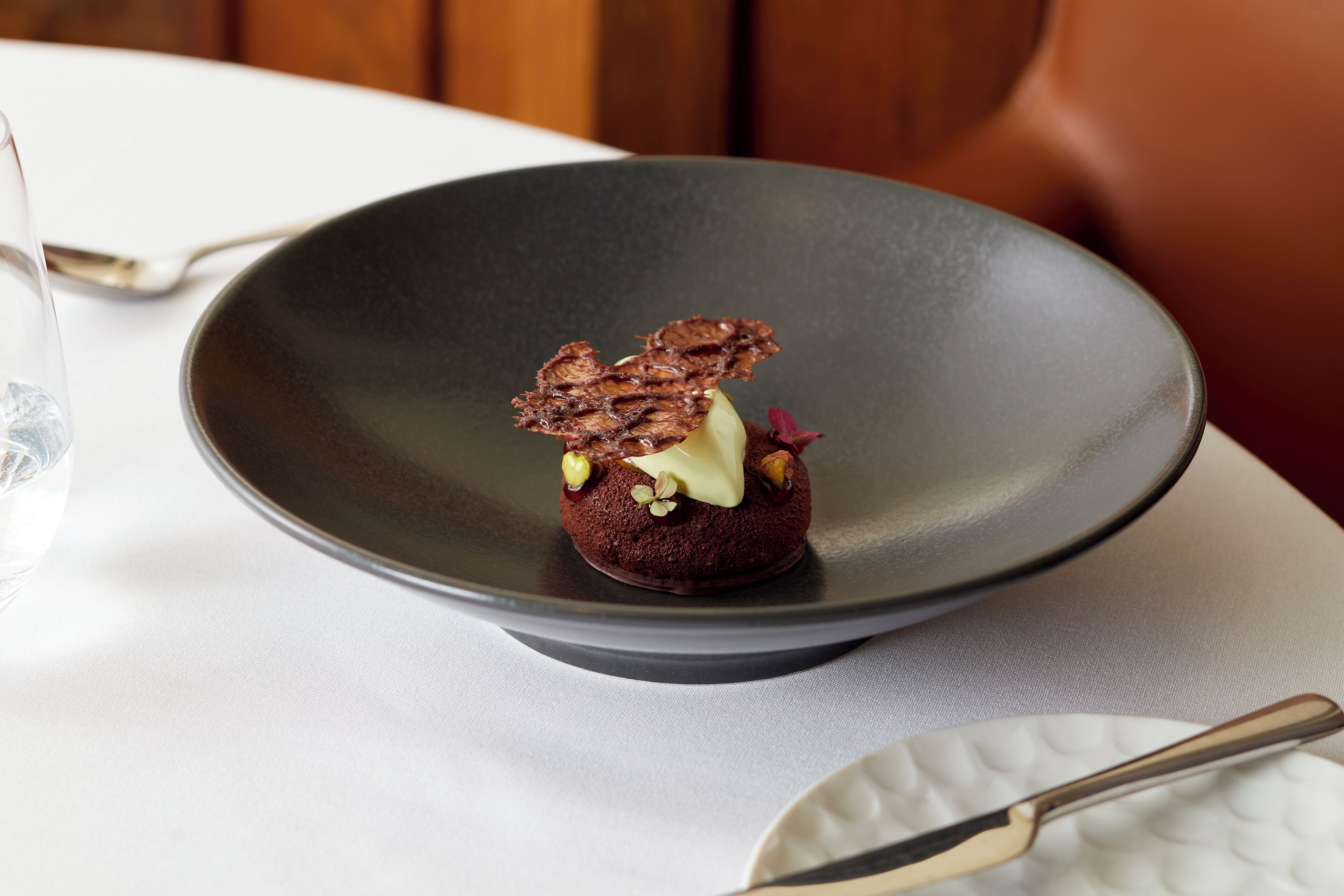 A dessert on a dark plate with a spoon and a glass of water on a table.