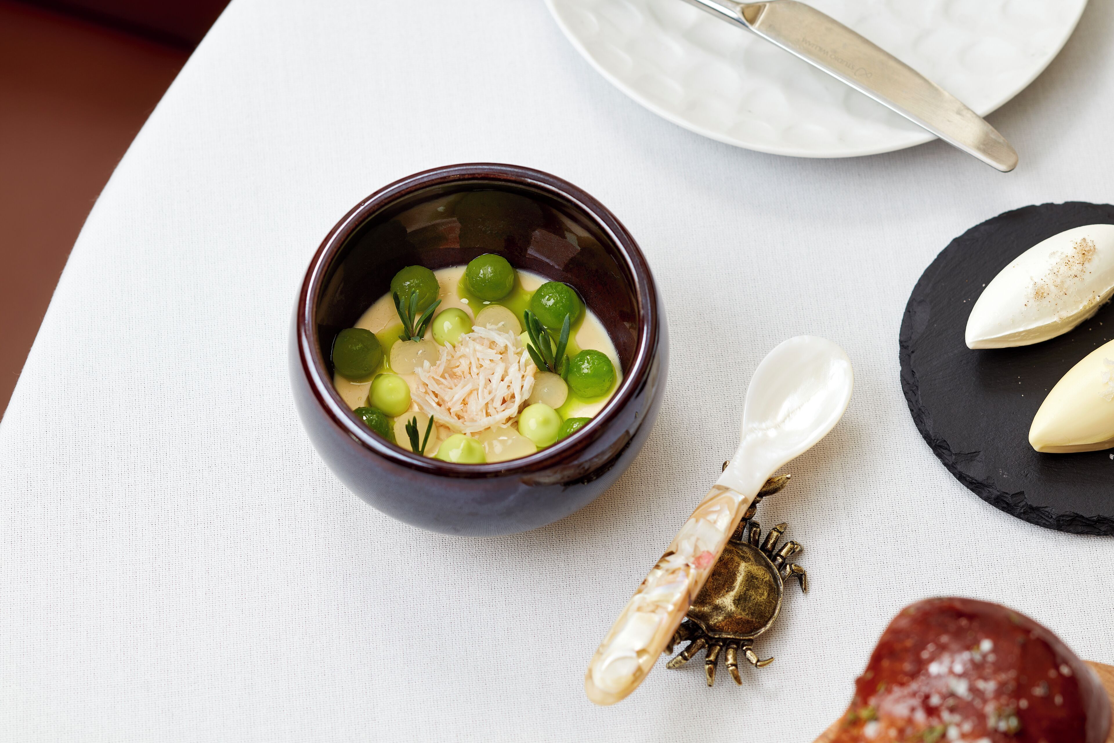 A bowl of soup with green vegetables and a decorative spoon on a white tablecloth.