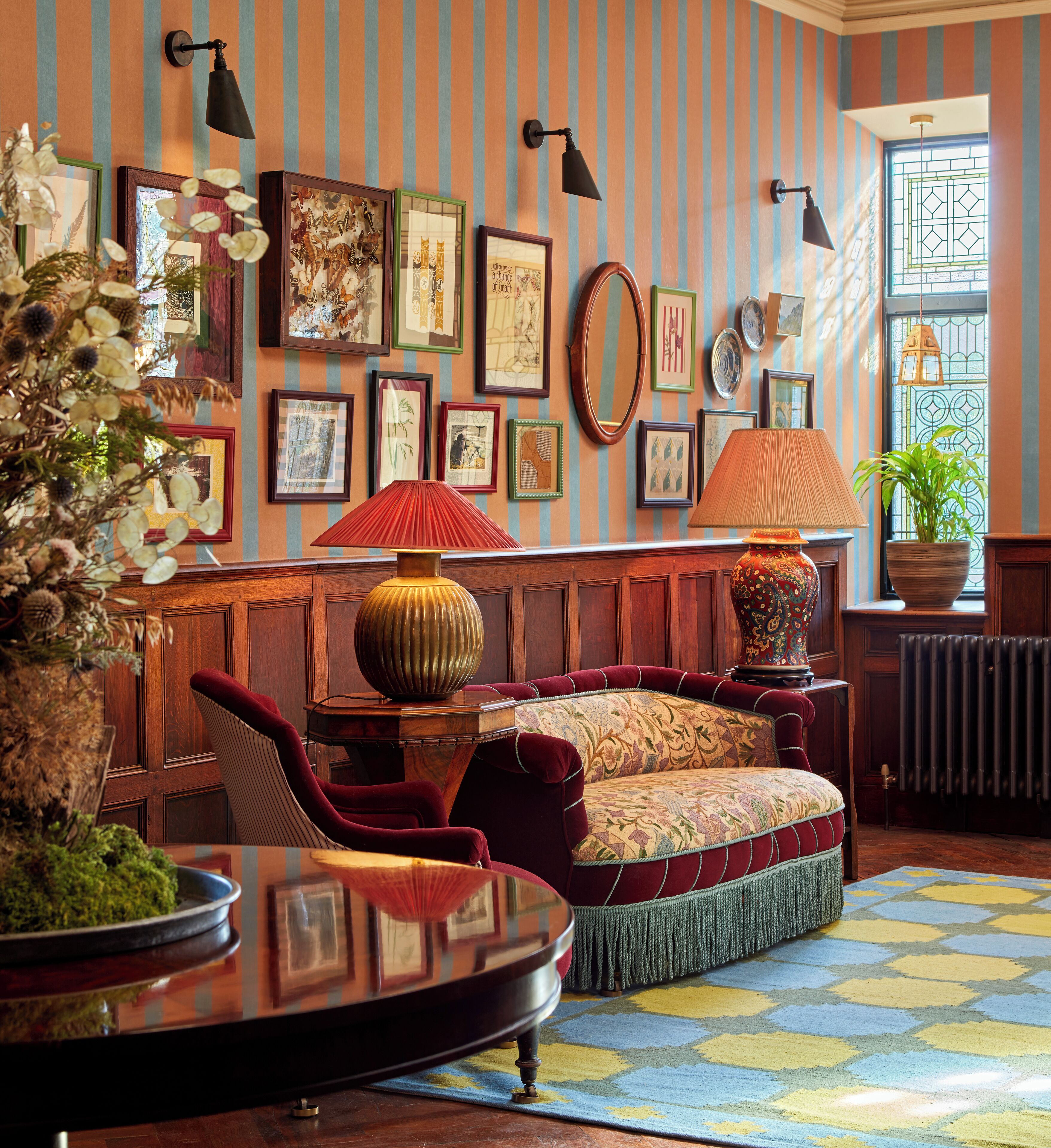A room with a patterned rug, a sofa with red cushions, a table with a lamp, and framed artwork on the wall.