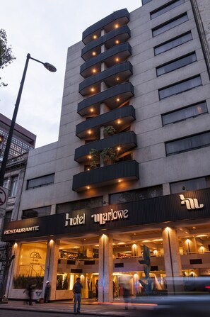 Un hôtel au design moderne, doté de nombreux balcons ornés de plantes, d’une entrée imposante et d’une enseigne de restaurant bien visible.