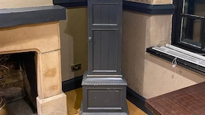 Un alto orologio a pendolo con cornice nera e dettagli dorati, posto in un angolo con una parete in pietra e una finestra alla destra.