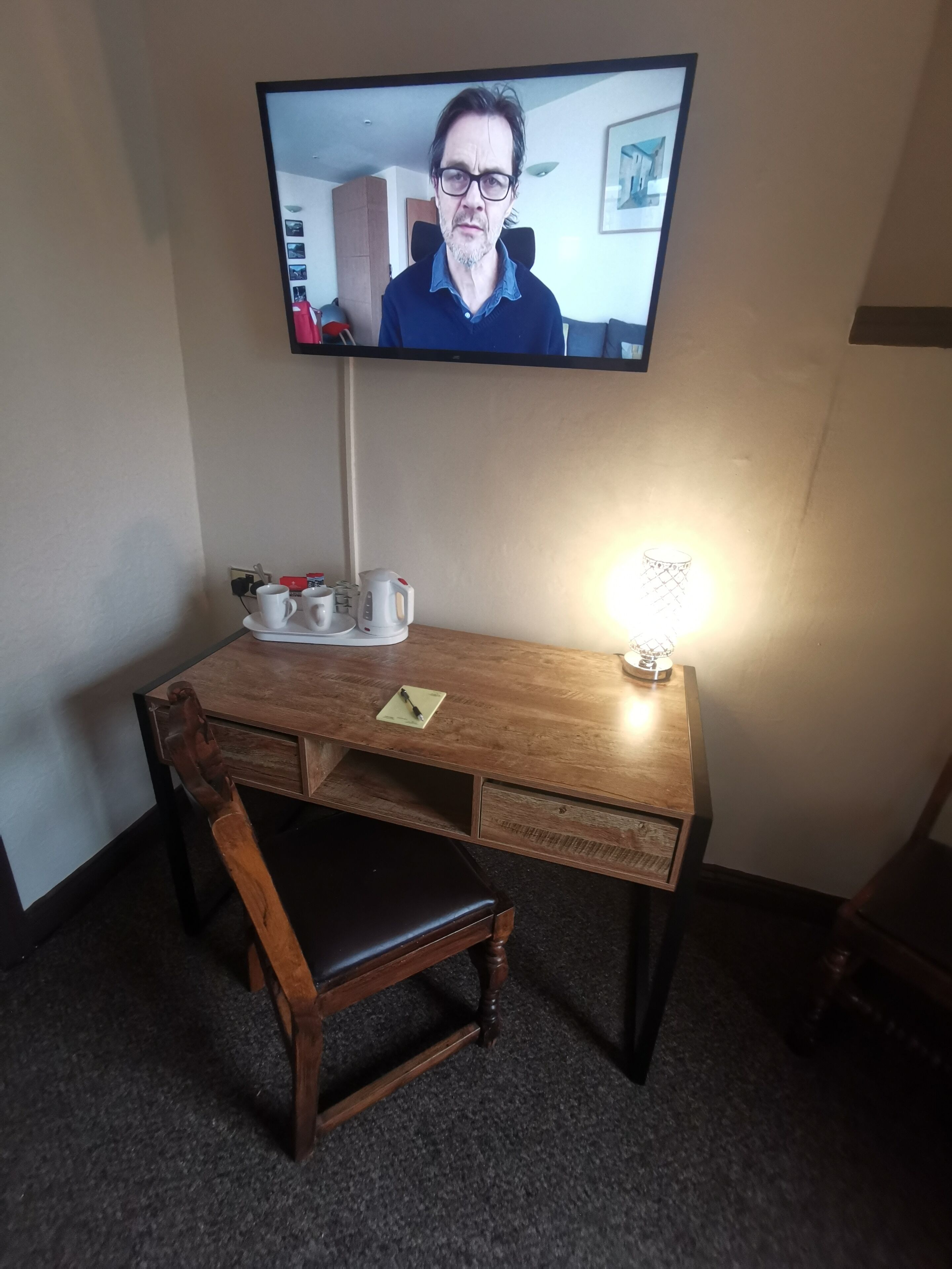 A hotel room with a wooden desk, a chair, a television mounted on the wall, and a lamp on the desk.