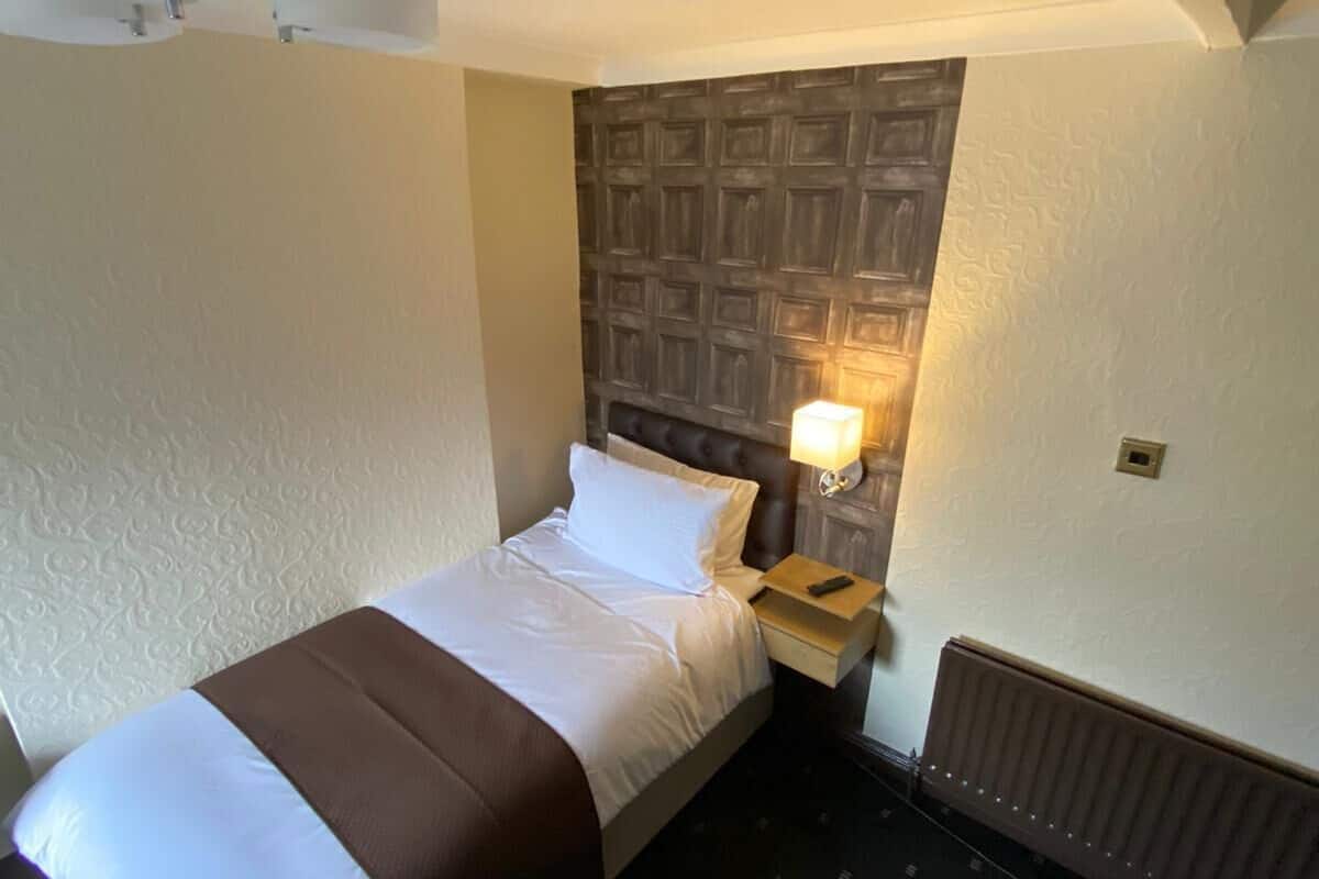 A hotel room with a bed, a nightstand, a wall-mounted lamp, and a radiator.