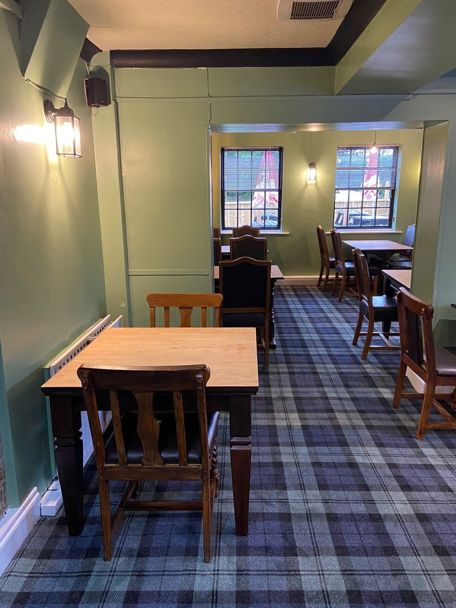 A dining area with plaid carpet, wooden tables and chairs, and two windows with blinds.