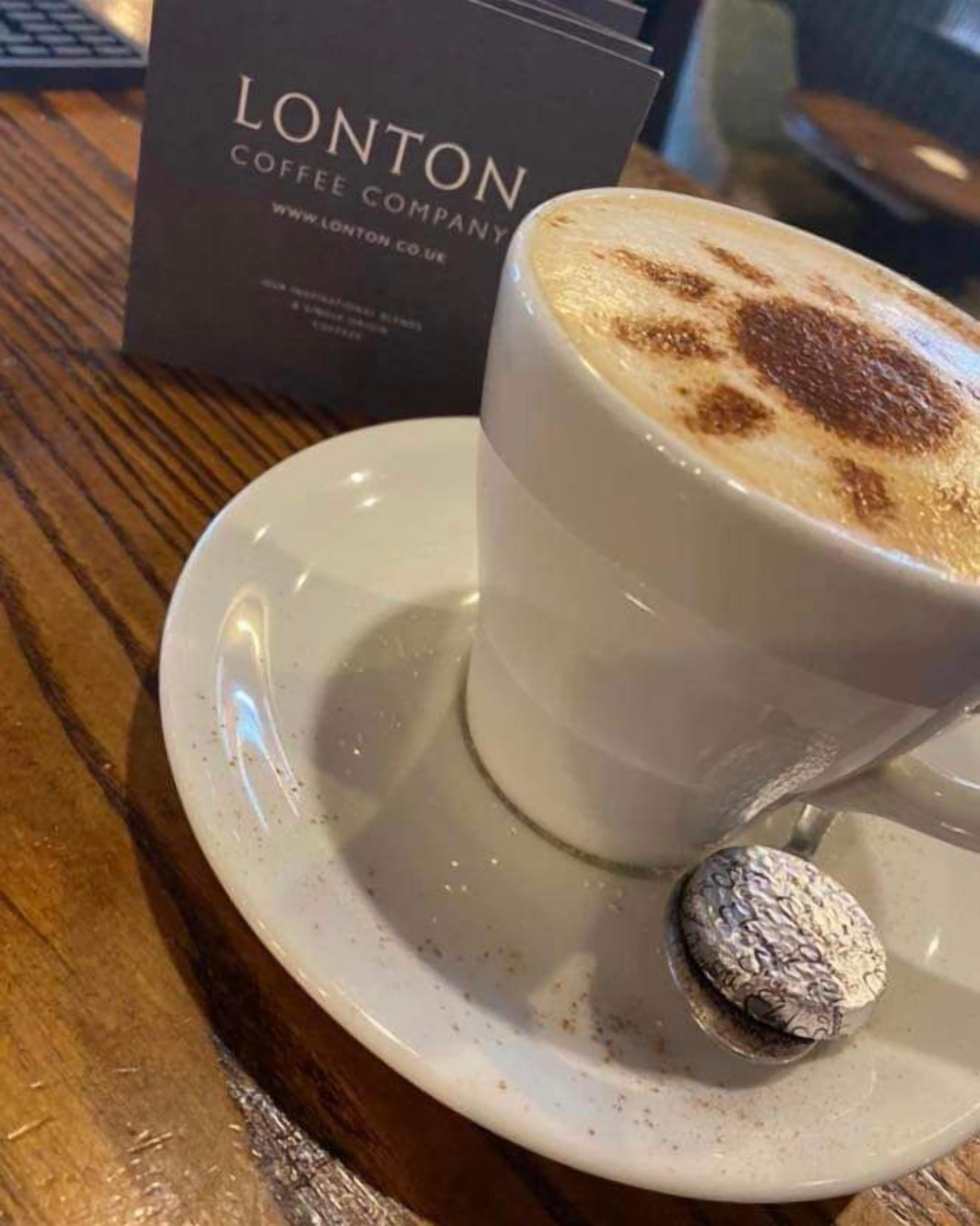 Une tasse de café surmontée d’une mousse onctueuse et accompagnée d’une boule de chocolat sur une soucoupe, le tout posé sur une table en bois, avec une enseigne de la société Lonton Coffee Company en arrière-plan.