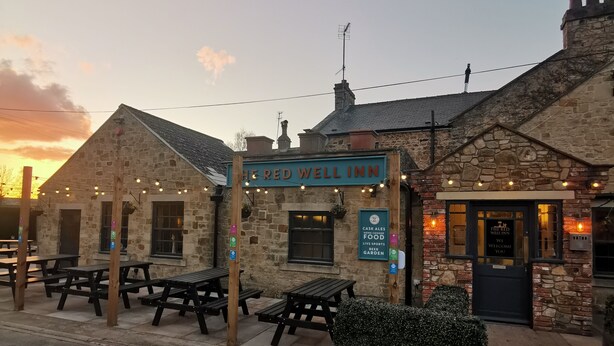 Un edificio in pietra con un'insegna che recita "The Red Well Inn" e posti a sedere all'aperto.