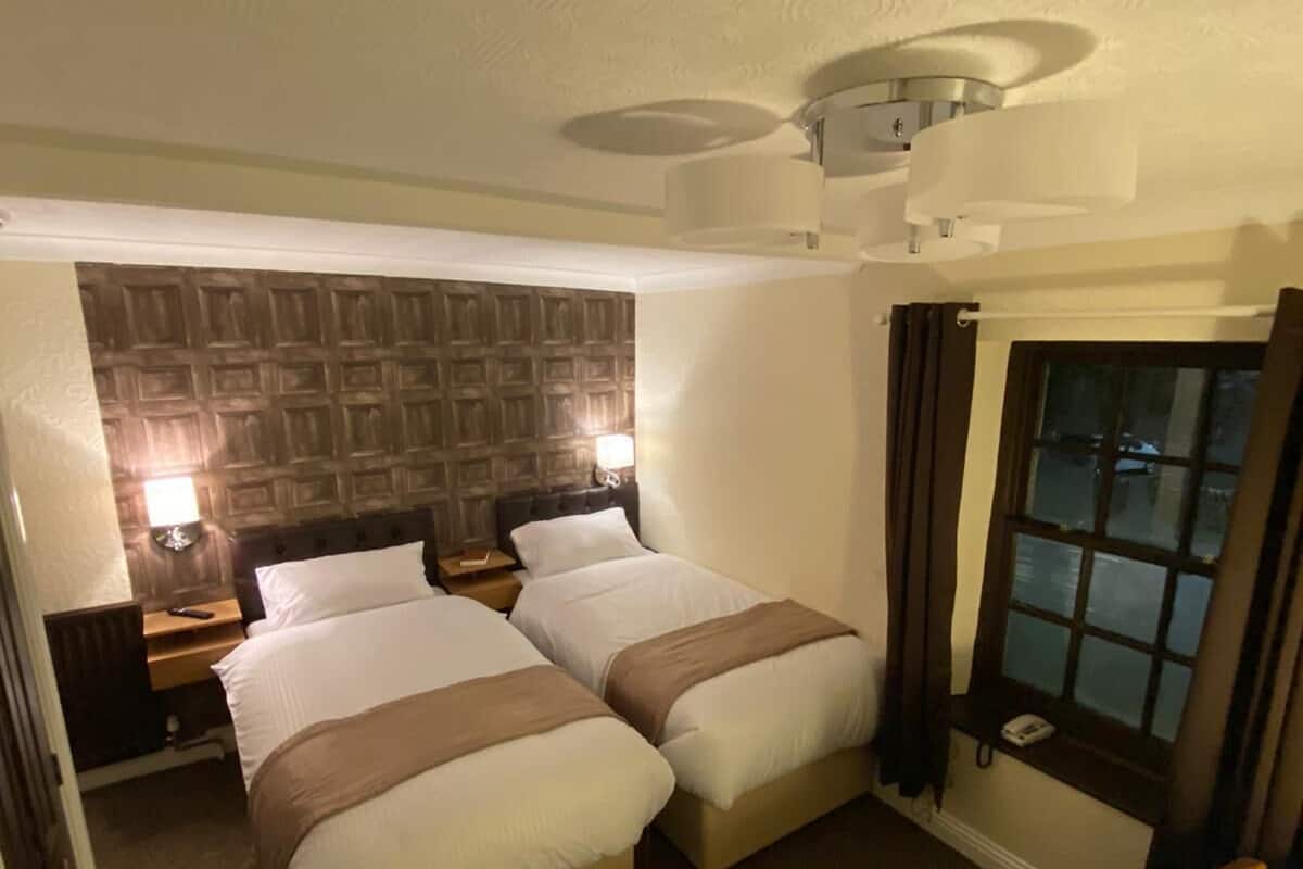 A hotel room with two beds, a wooden headboard, a small window with curtains, and a ceiling light fixture.