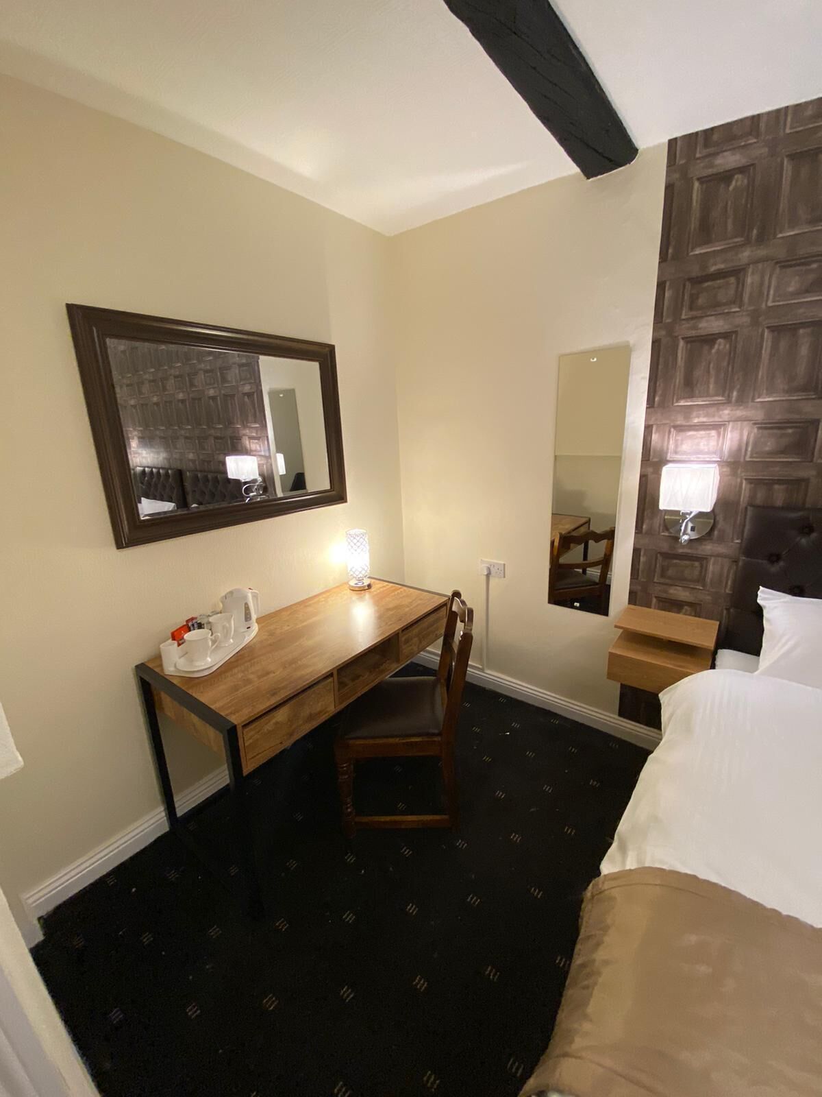 A hotel room with a wooden desk, a chair, a bed, a nightstand, a mirror, and a wall-mounted light.