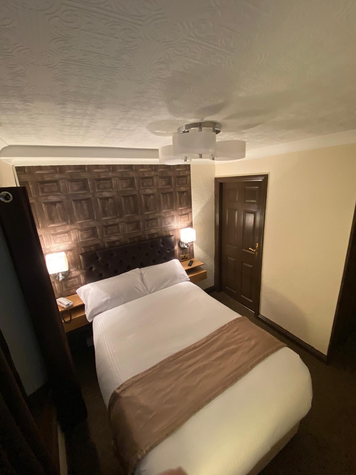 A hotel room with a bed, a wooden headboard, a nightstand with a lamp, a door, and a ceiling light fixture.