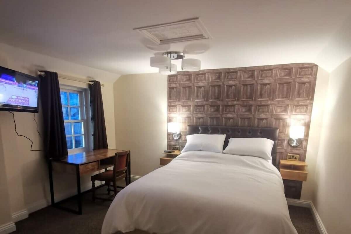 A hotel room with a bed, a desk, a chair, a TV, and a wall-mounted light fixture.