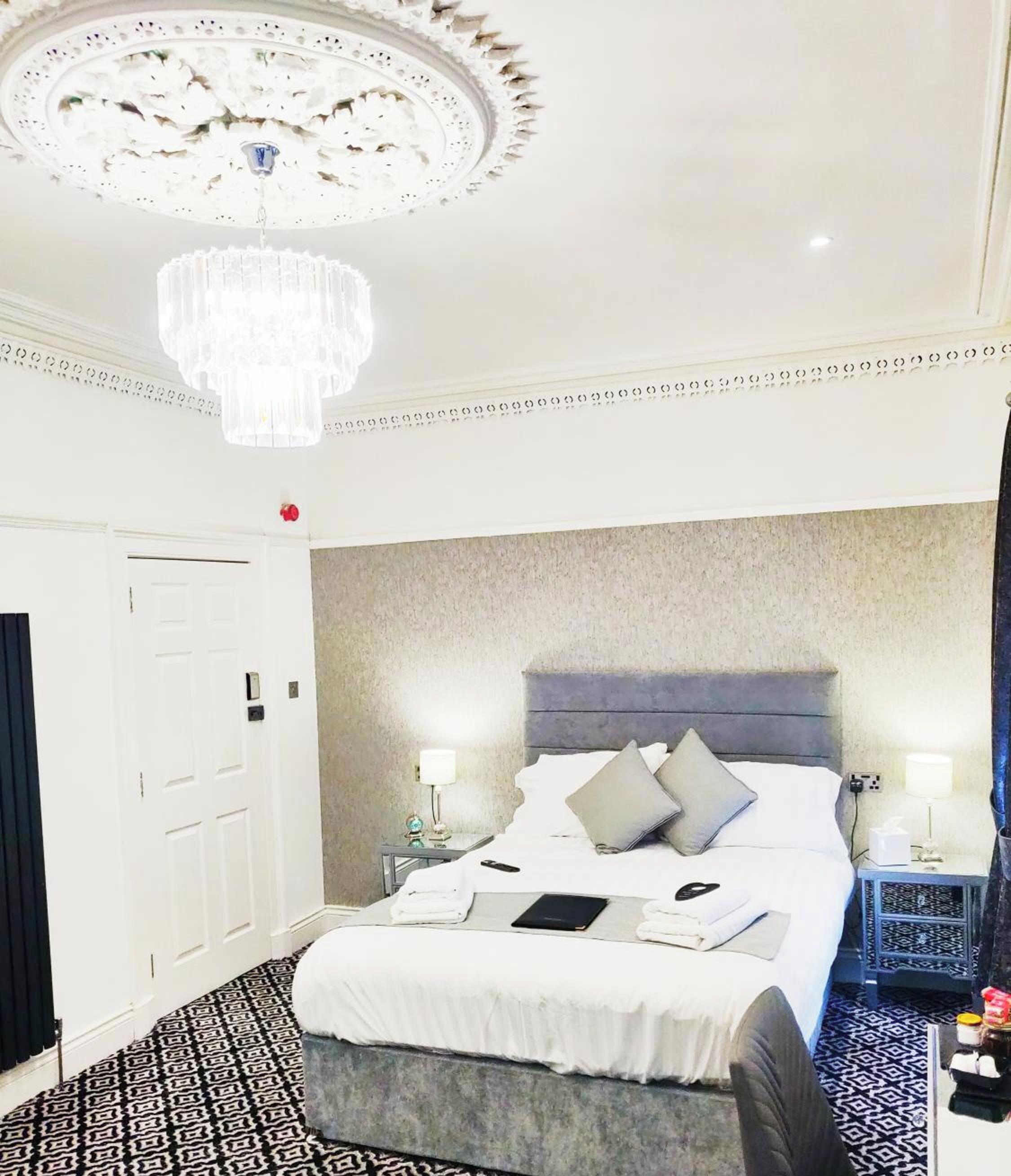 A hotel room with a large bed, a chandelier, a bedside table with a lamp, and a patterned floor.